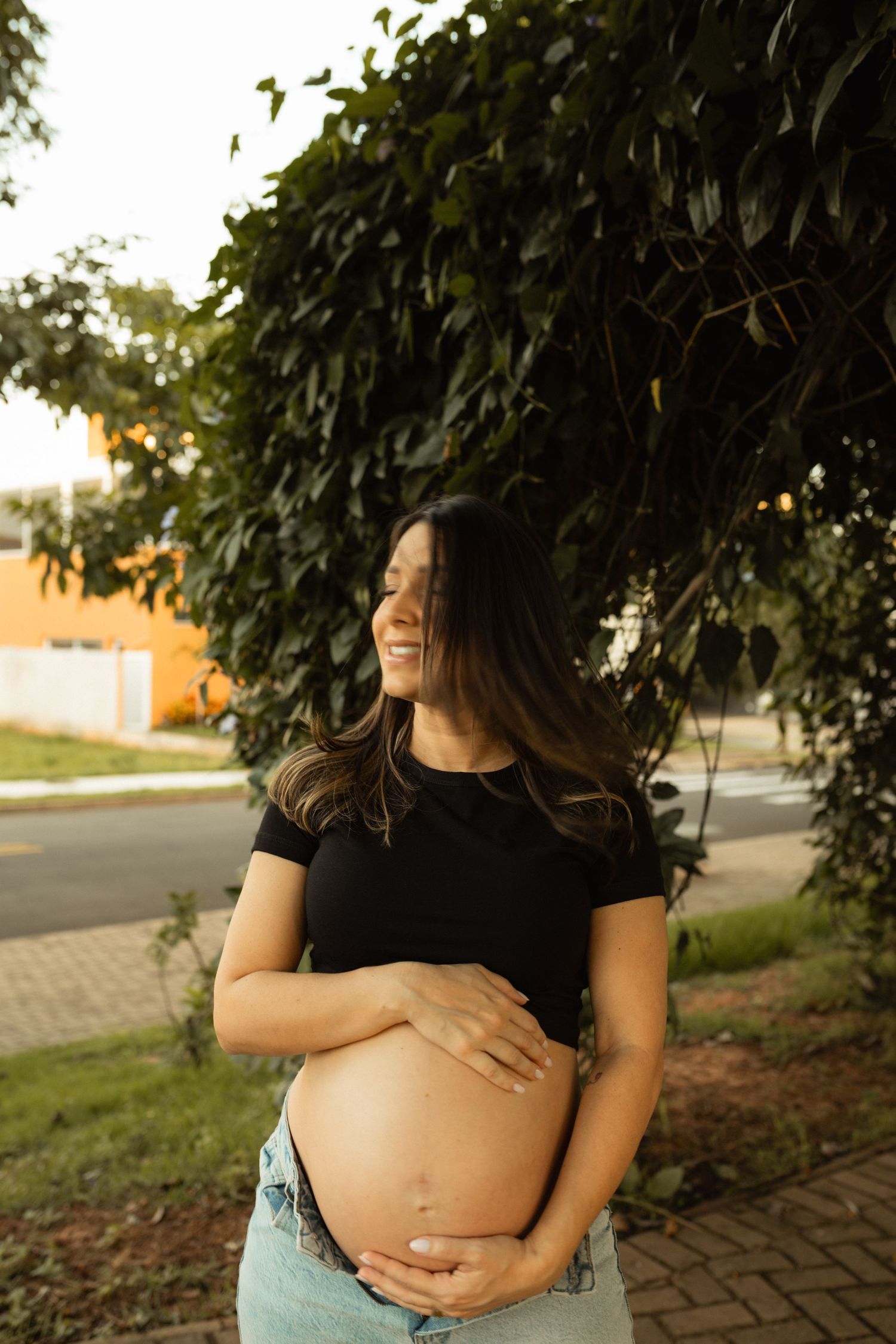 Smiling pregnant woman in black top joyfully cradling her baby bump under lush green foliage outdoors.
