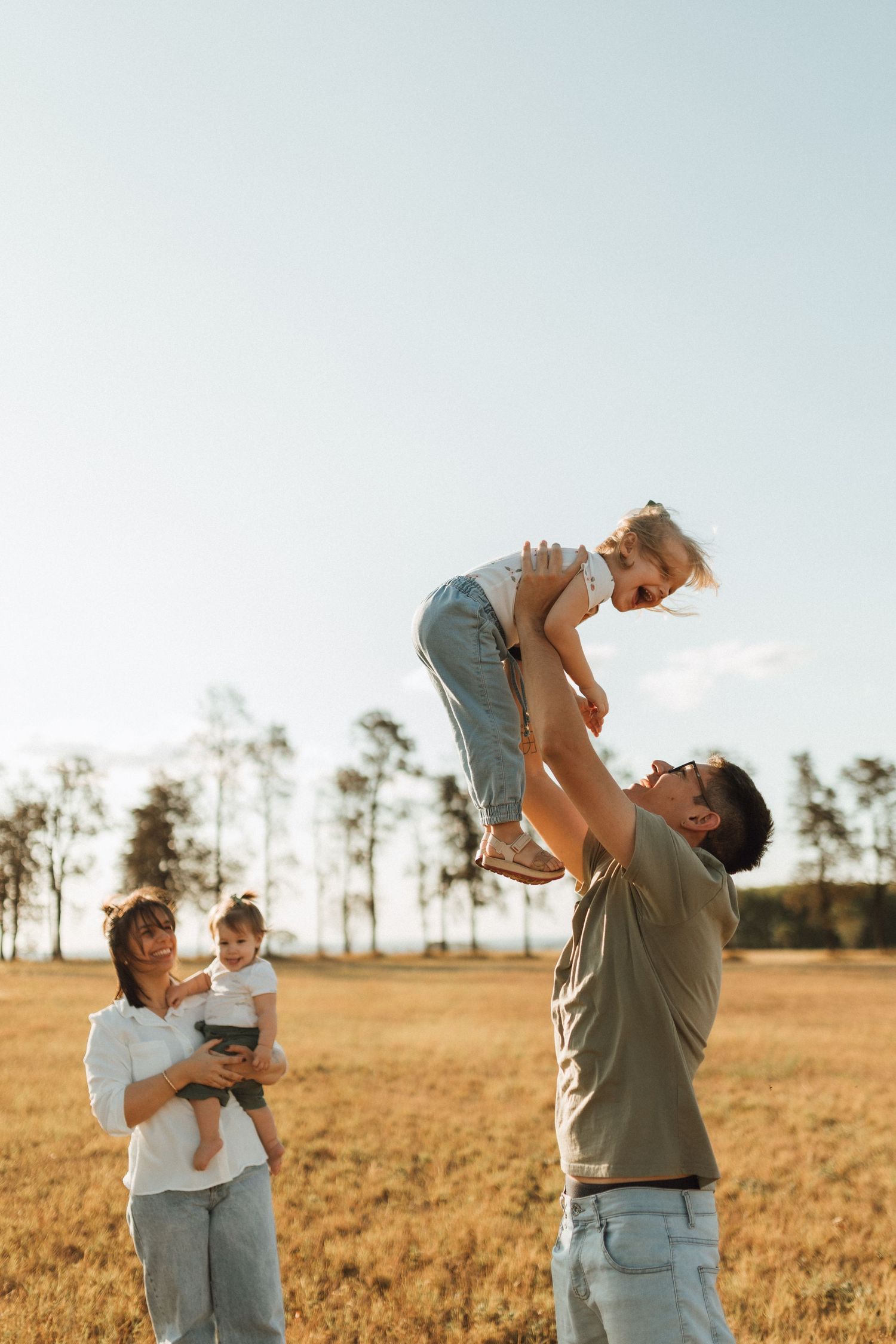 Father joyfully lifts child in air while mother holds baby in sunny field.