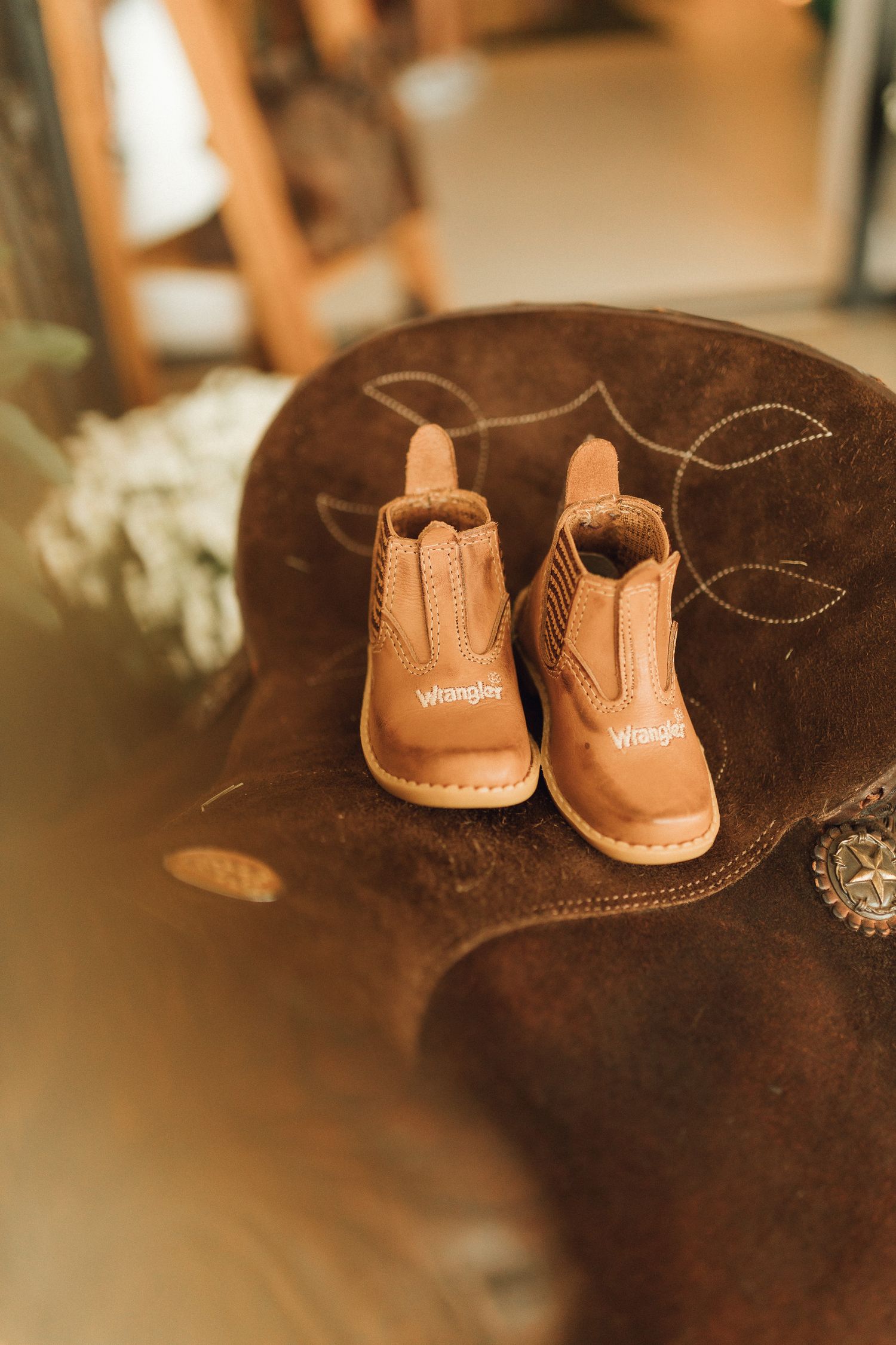 Tiny Wrangler brand tan leather baby cowboy boots sitting on a brown suede cowboy hat with decorative stitching.