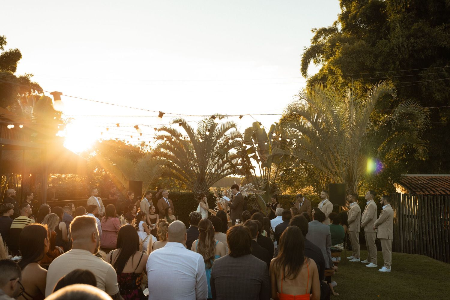 Outdoor wedding ceremony at golden hour with guests gathered on a lawn surrounded by palm trees and string lights.
