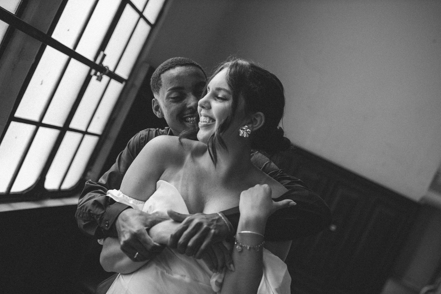 Black and white photo of a groom hugging bride from behind, both smiling joyfully near a window.