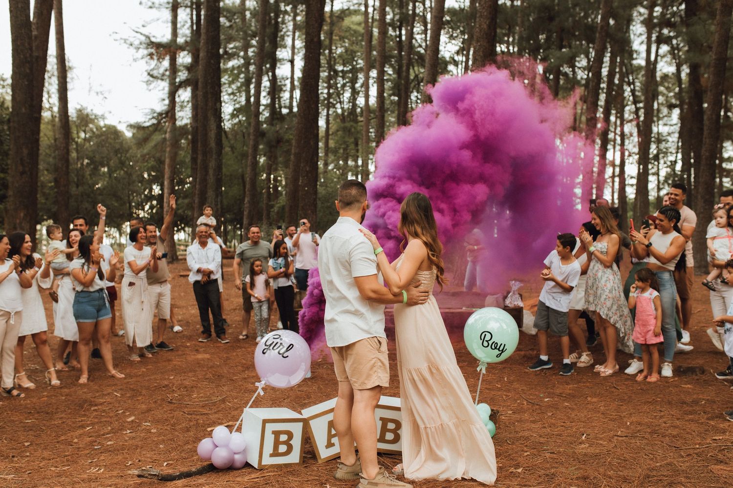 Couple reveals baby gender with purple smoke bomb surrounded by cheering guests in a pine forest.