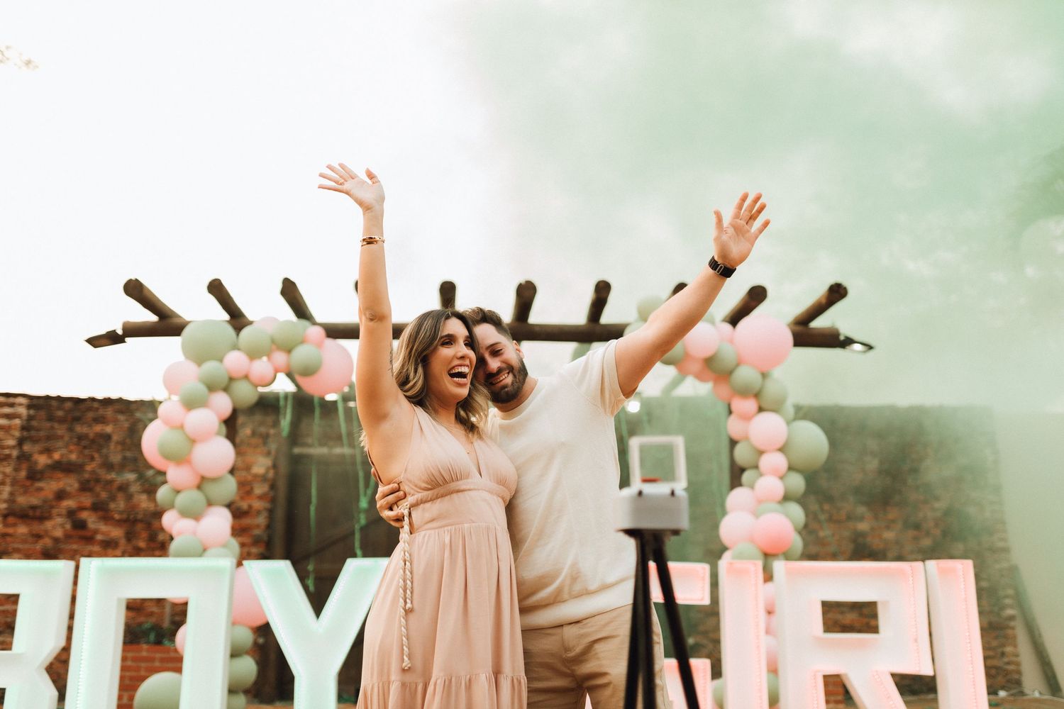 Joyful couple celebrating with raised arms at outdoor gender reveal party with colorful smoke.