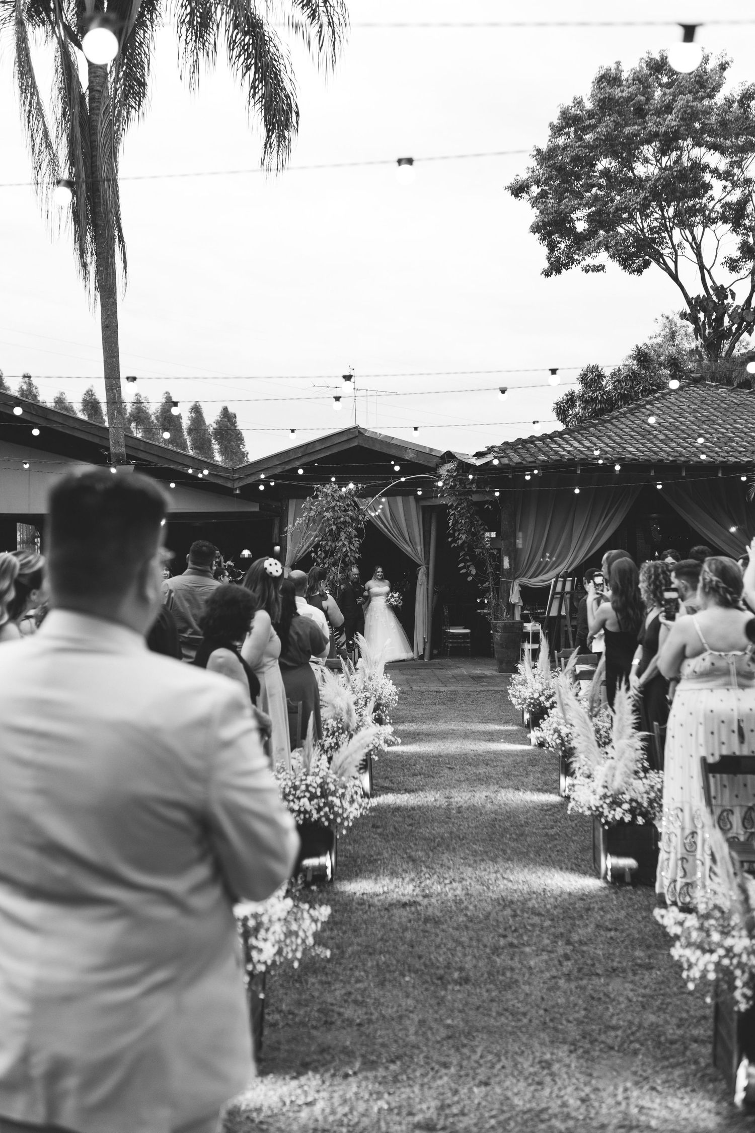 Black and white outdoor wedding ceremony with bride walking down flower-lined aisle.