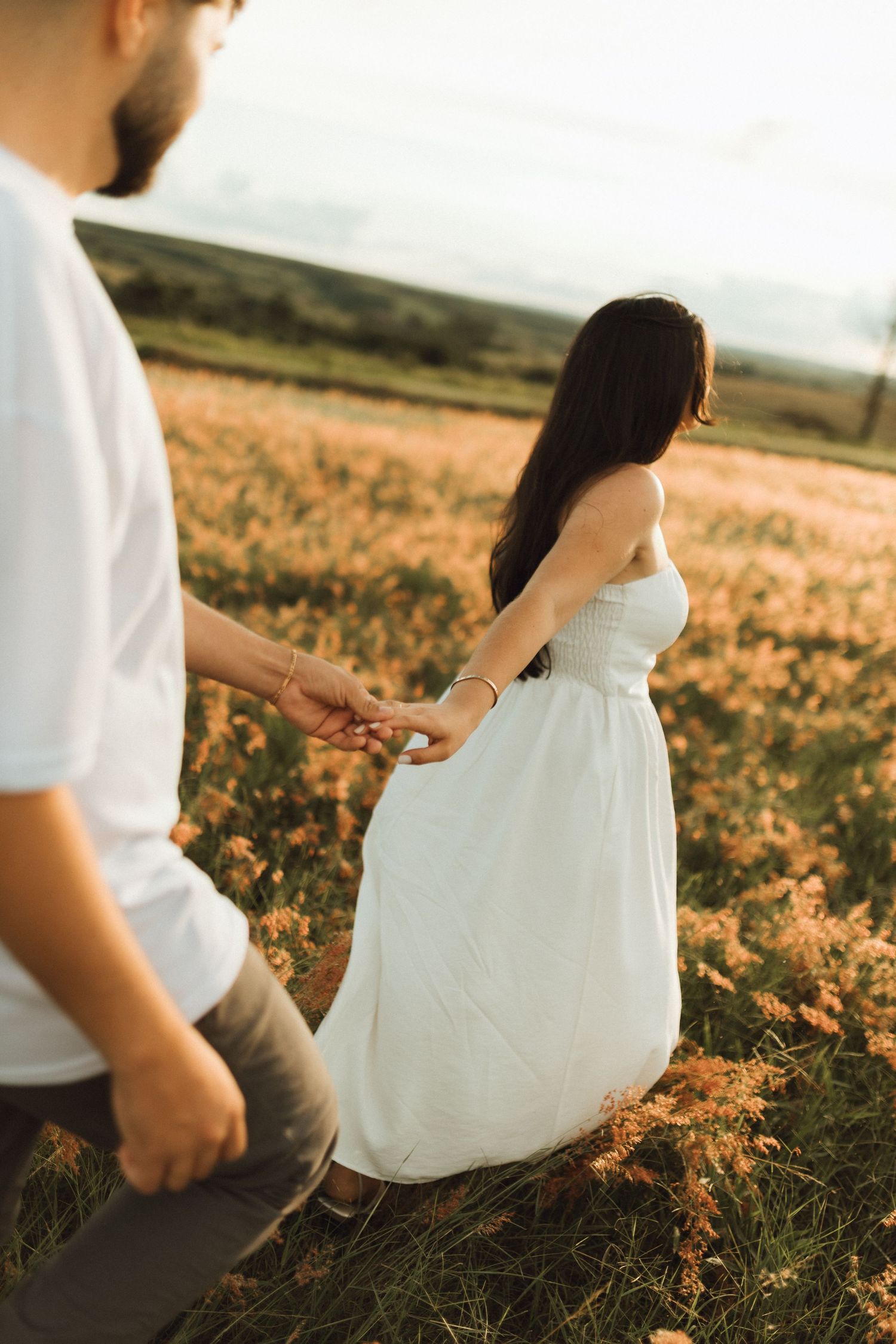 Couple holding hands walking through a golden flower field at sunset in white outfits.