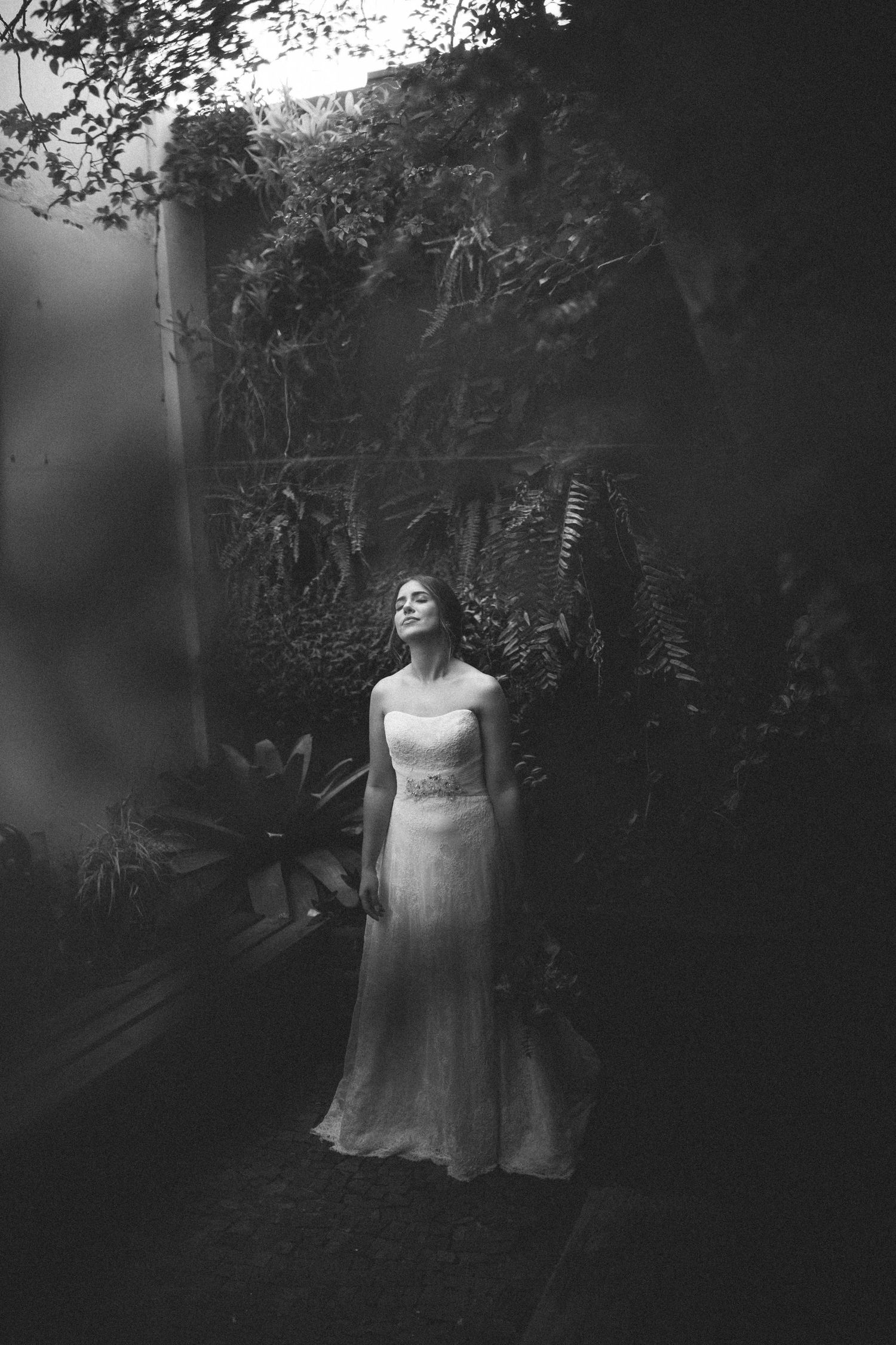 Elegant bride in lace gown poses with eyes closed among lush tropical plants in black and white.