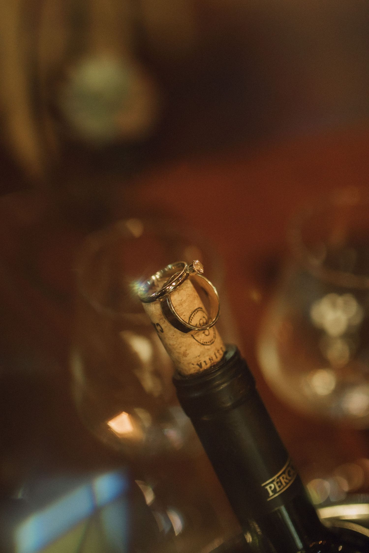 Wedding rings resting on a wine bottle cork with blurred glasses in background.