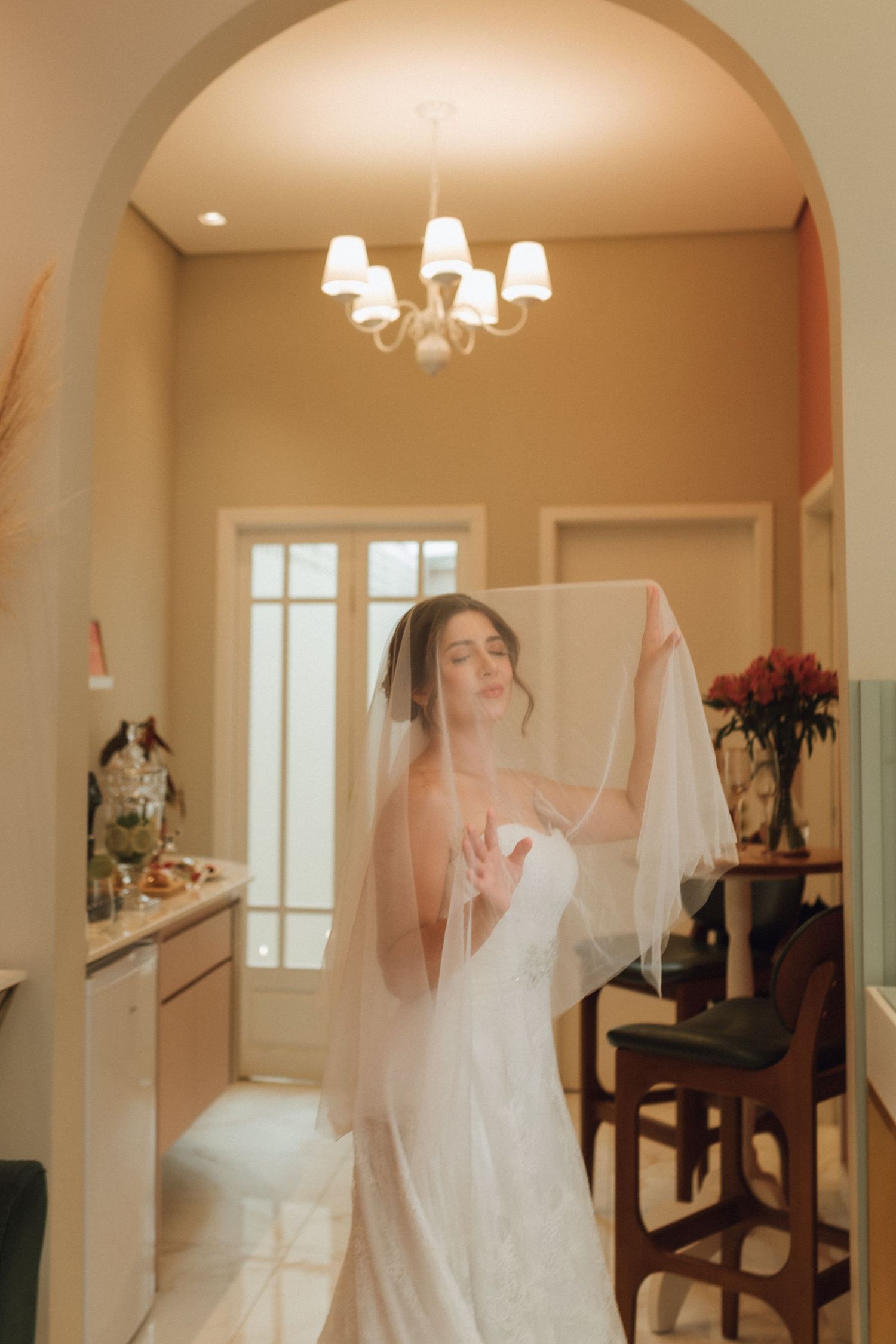 Elegant bride in white gown lifting veil under chandelier in warmly lit arched interior room.