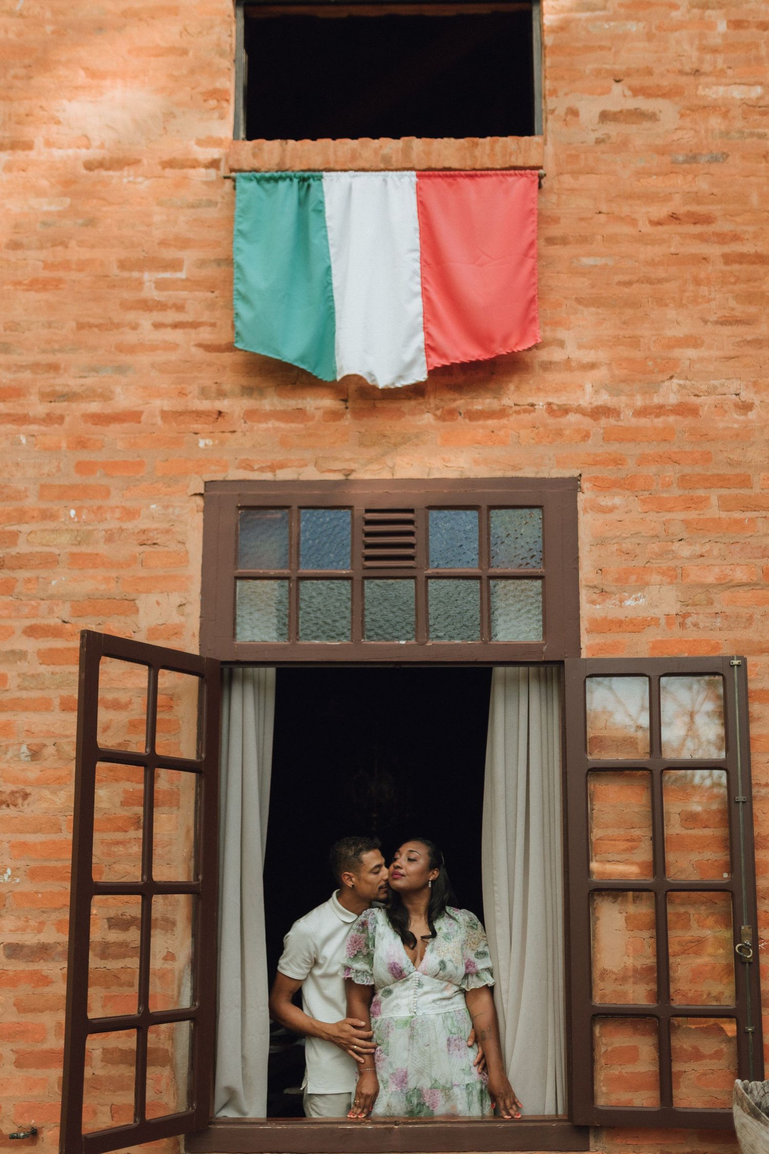 Couple sharing a tender kiss in an open window with Italian flag above.