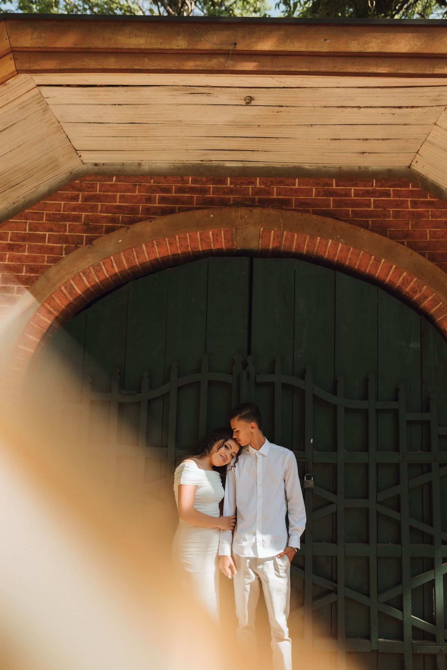 Couple leaning together affectionately framed by brick archway and vintage wooden door outdoors.
