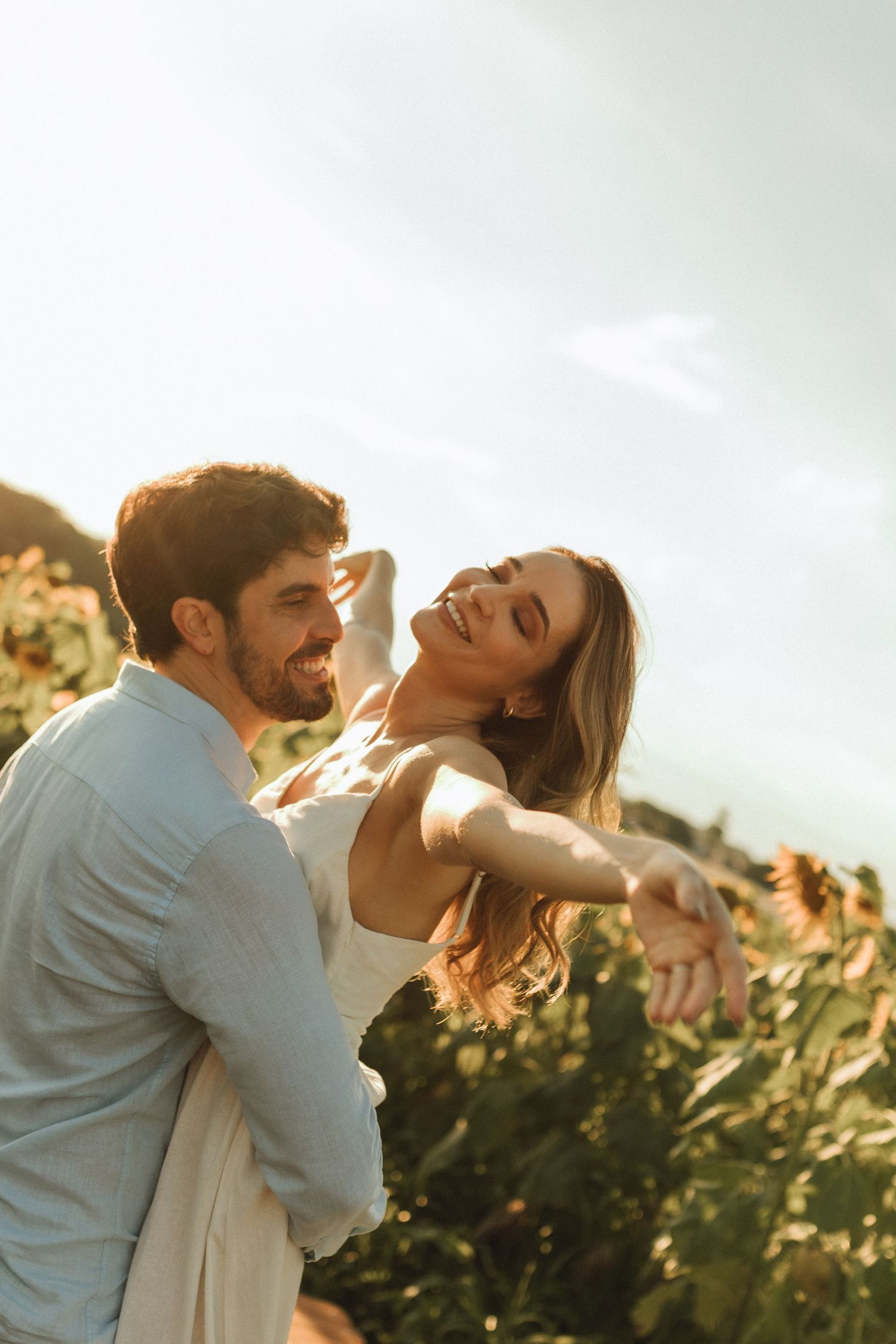 Joyful couple dancing and laughing together in sunflower field during golden hour sunset.
