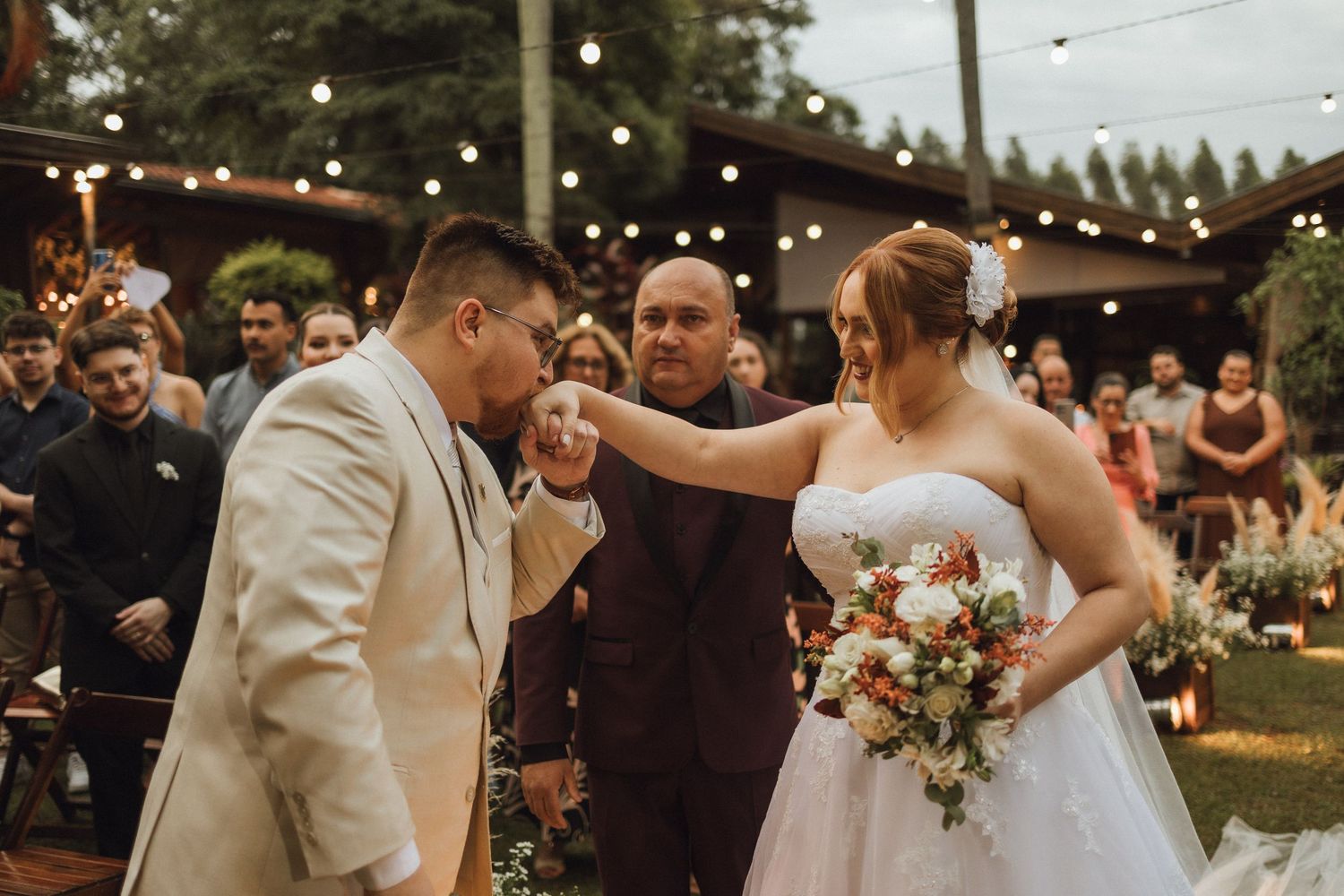 Groom in beige suit holds brides hand during outdoor wedding ceremony with string lights.