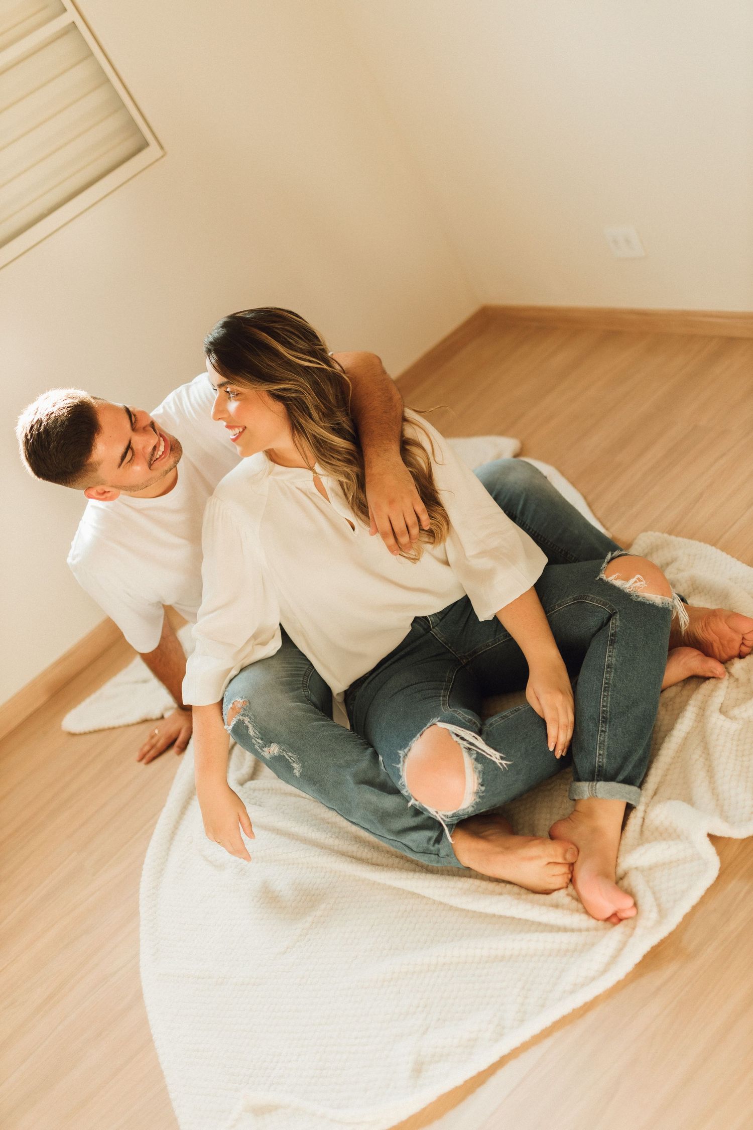 Happy couple sitting on a blanket on the floor, wearing white tops and jeans, sharing a tender moment.