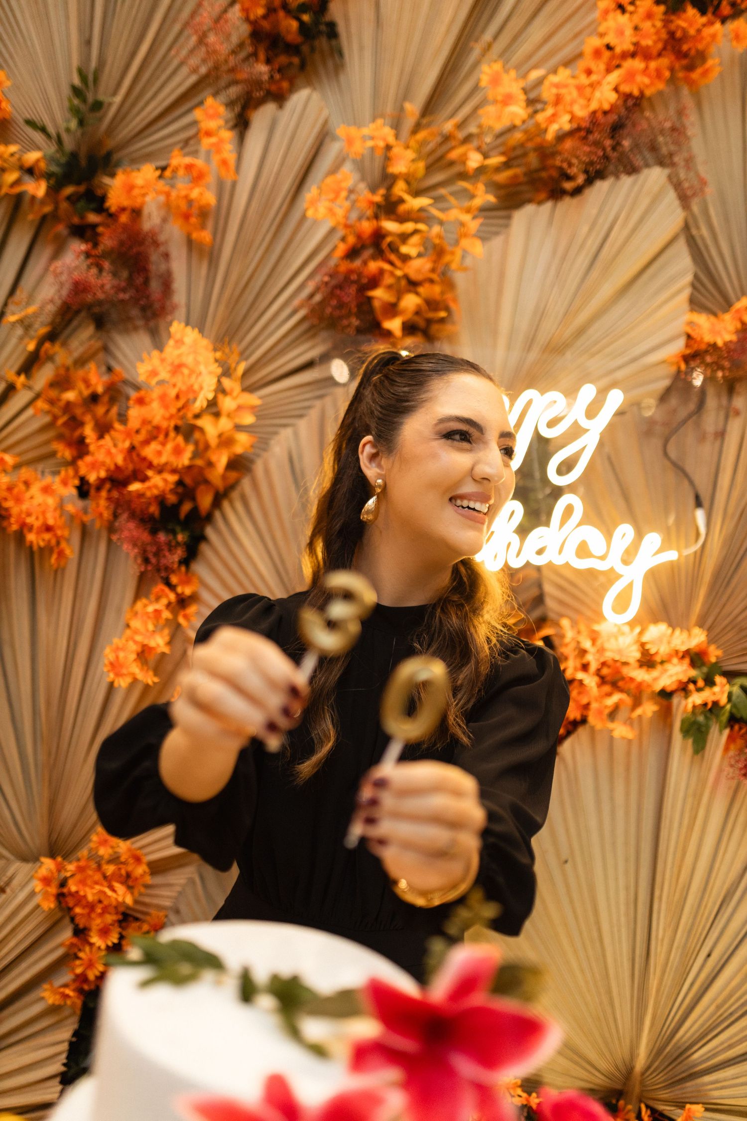 Woman celebrating 30th birthday holding gold number candles before white cake with orange floral backdrop and neon happy birthday sign.