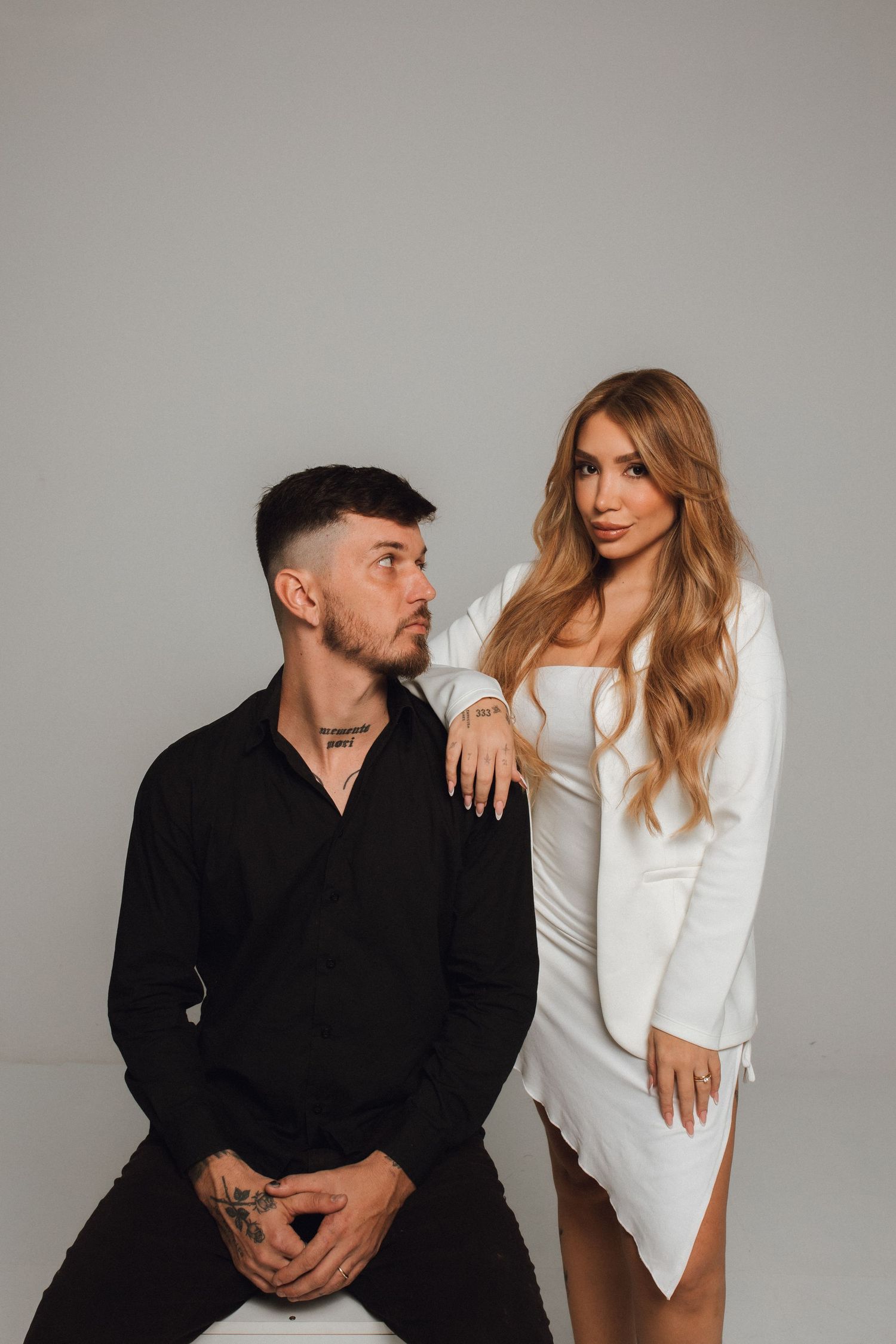 A tattooed man in black shirt sits beside a blonde woman in white dress, posing together against a gray background.