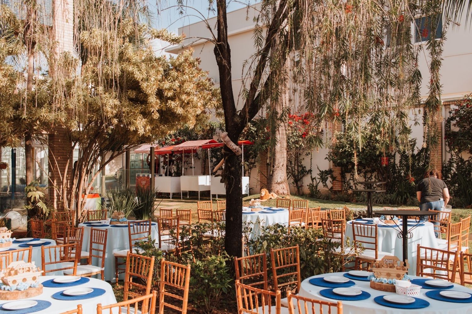 Outdoor wedding reception with wooden chiavari chairs, blue-accented table settings, and lush trees in a sunlit courtyard.