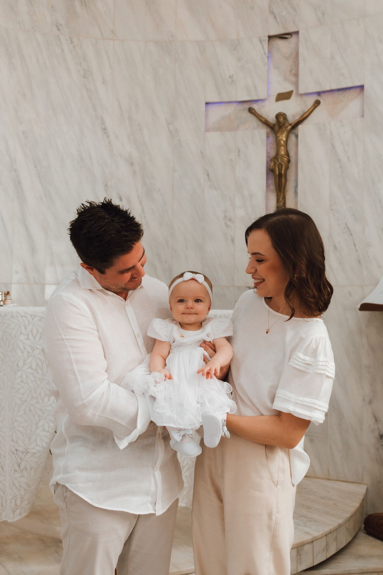 Family celebrating baby baptism in white outfits before a marble altar with crucifix in a church.