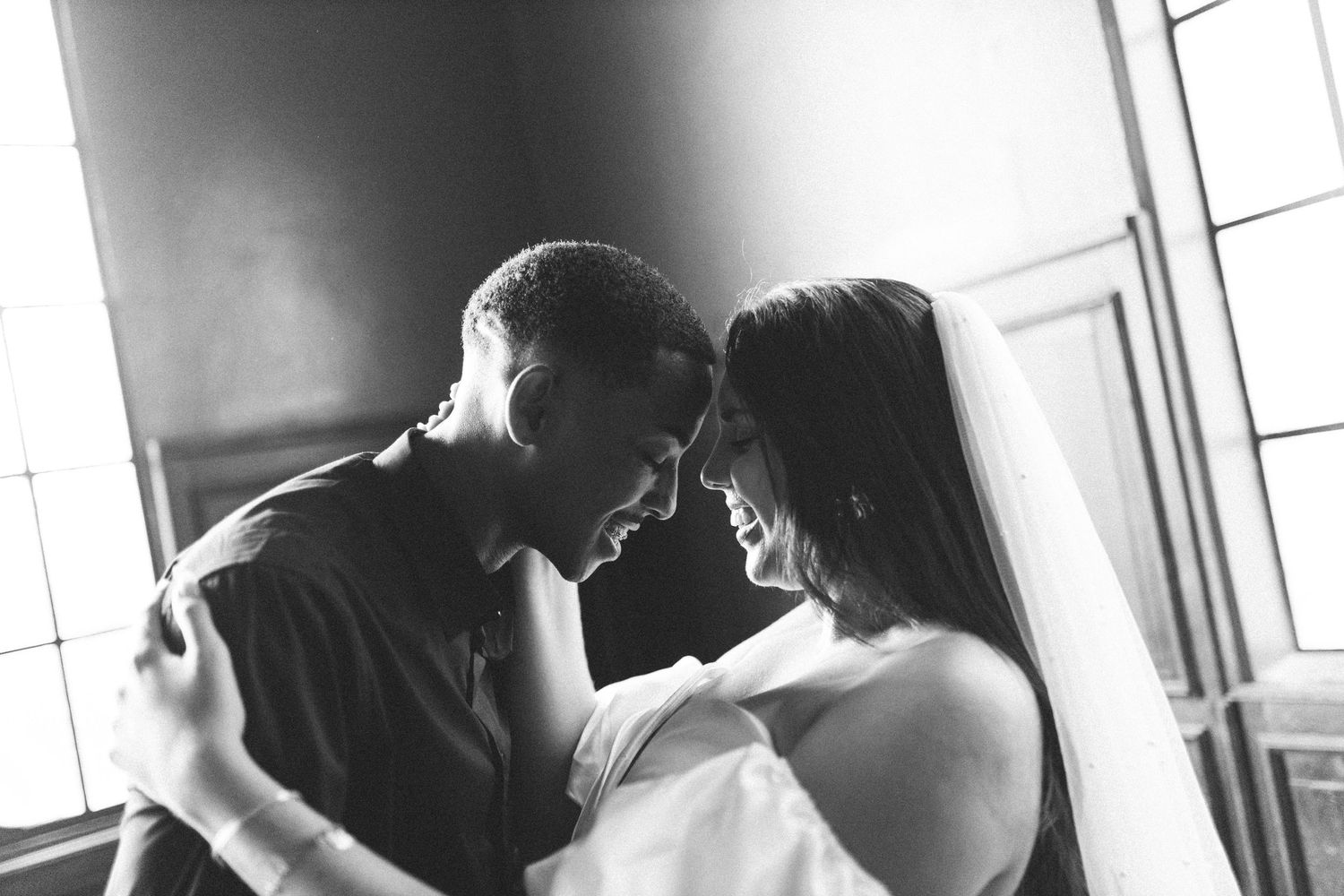 Black and white photo of a bride and groom sharing an intimate moment, touching foreheads and smiling at each other.