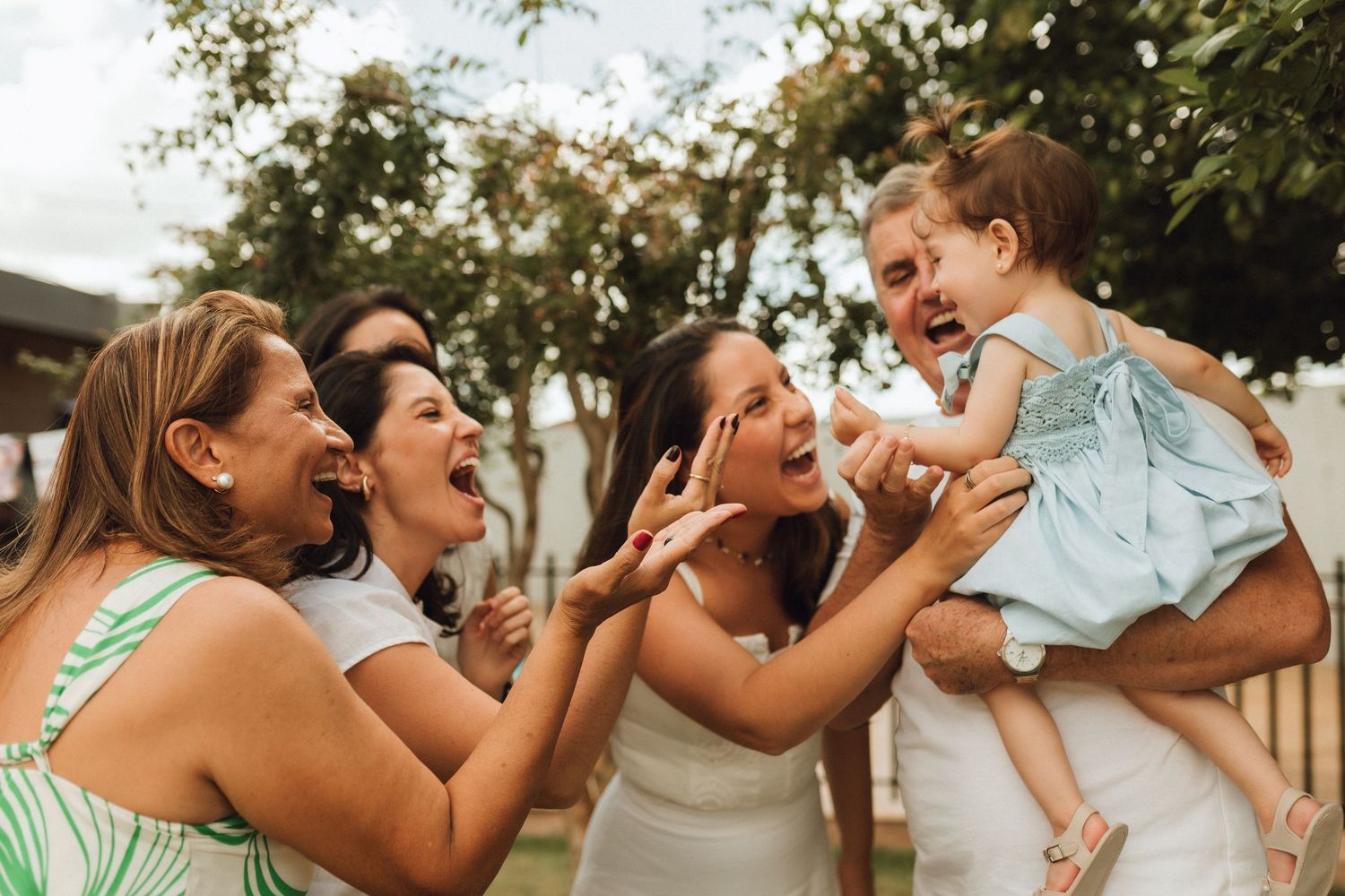 A joyful family group laughing and celebrating outdoors, with a woman holding a smiling toddler in a blue dress.