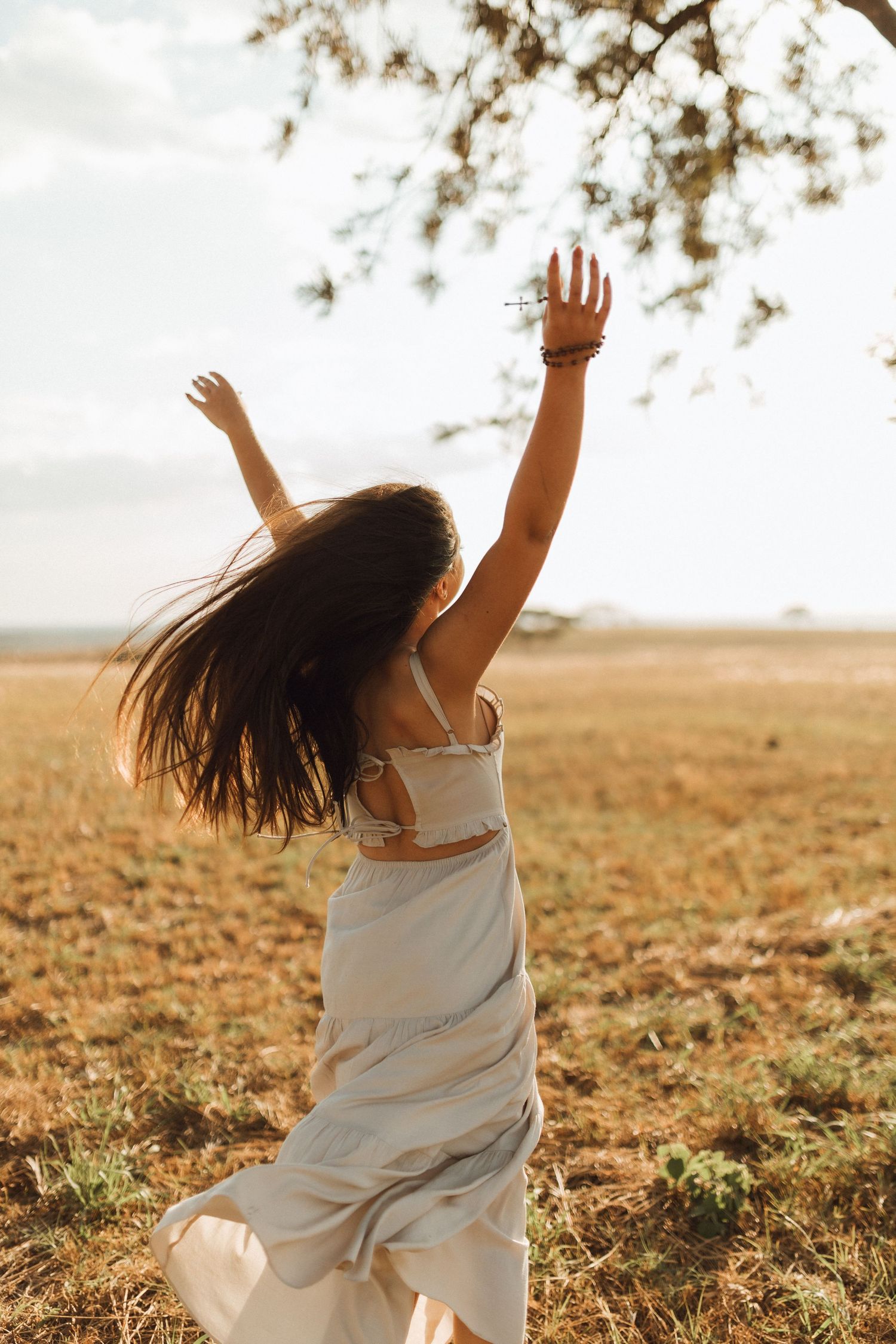 Carefree young woman in white dress dancing in golden field with arms raised toward tree branches.
