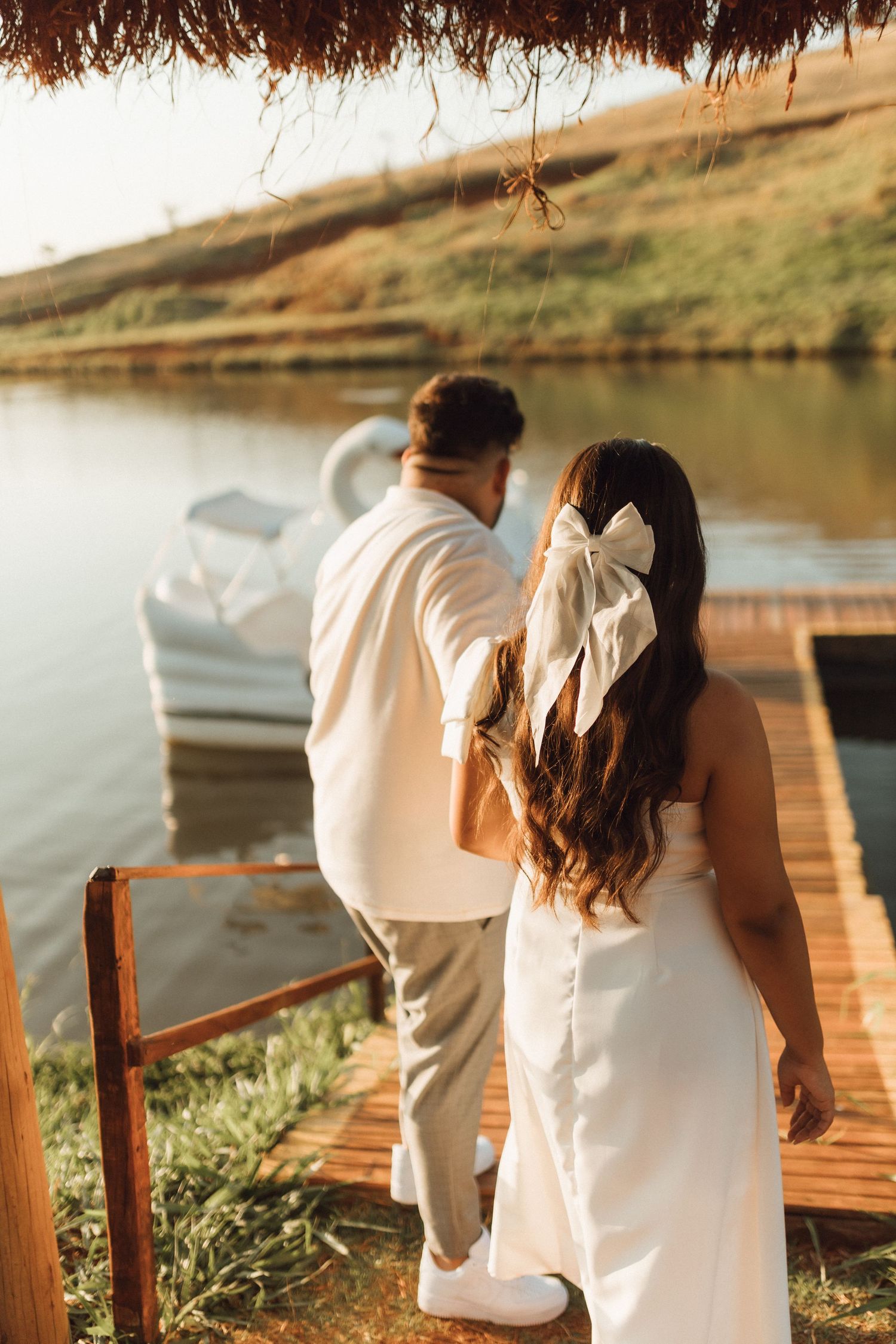 Bride and groom in white attire stand on wooden dock by calm lake at golden hour, woman wearing large bow hair accessory.