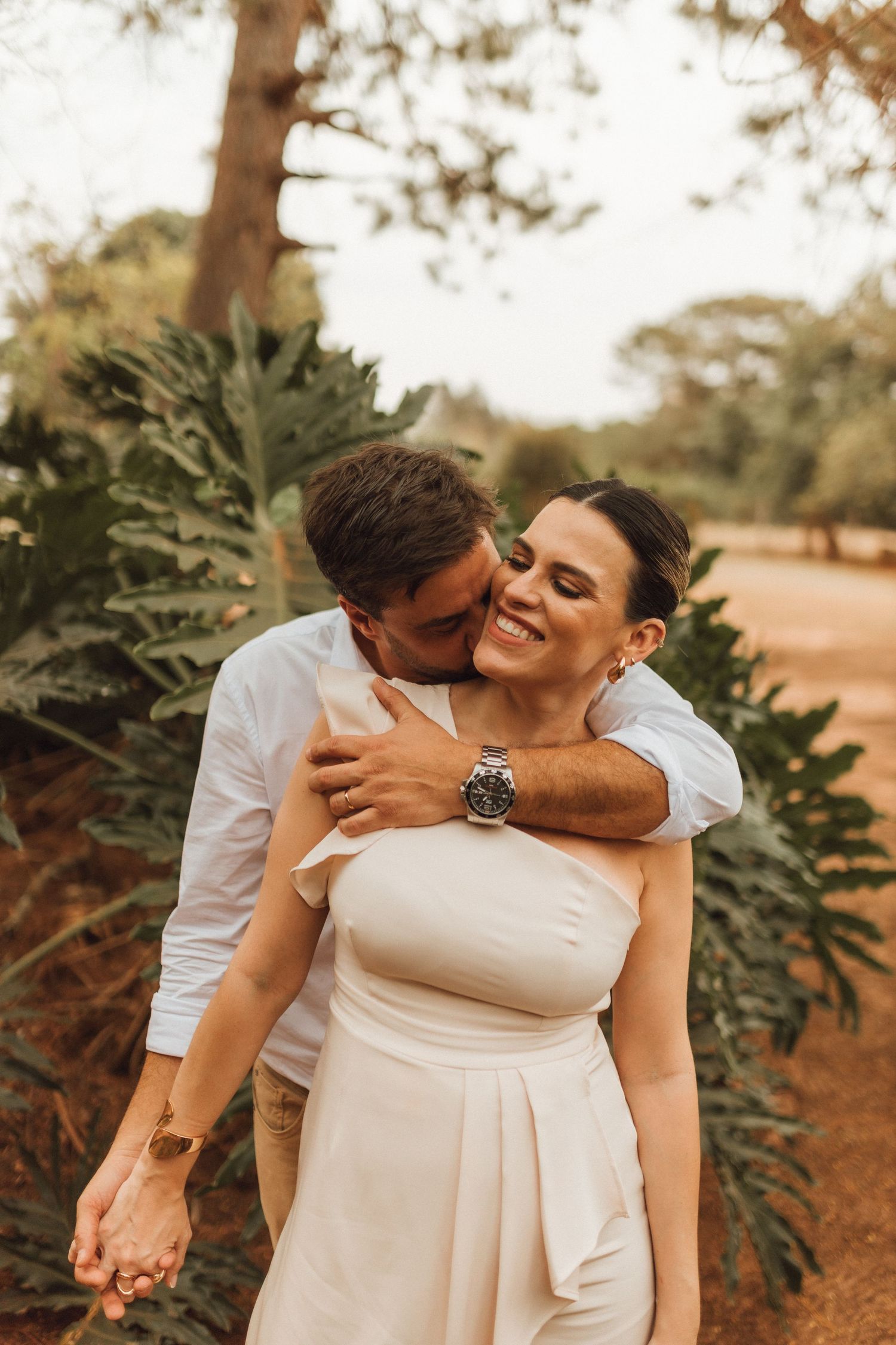 Couple embracing outdoors, groom kissing bride on cheek, bride smiling in white dress, lush green garden background.