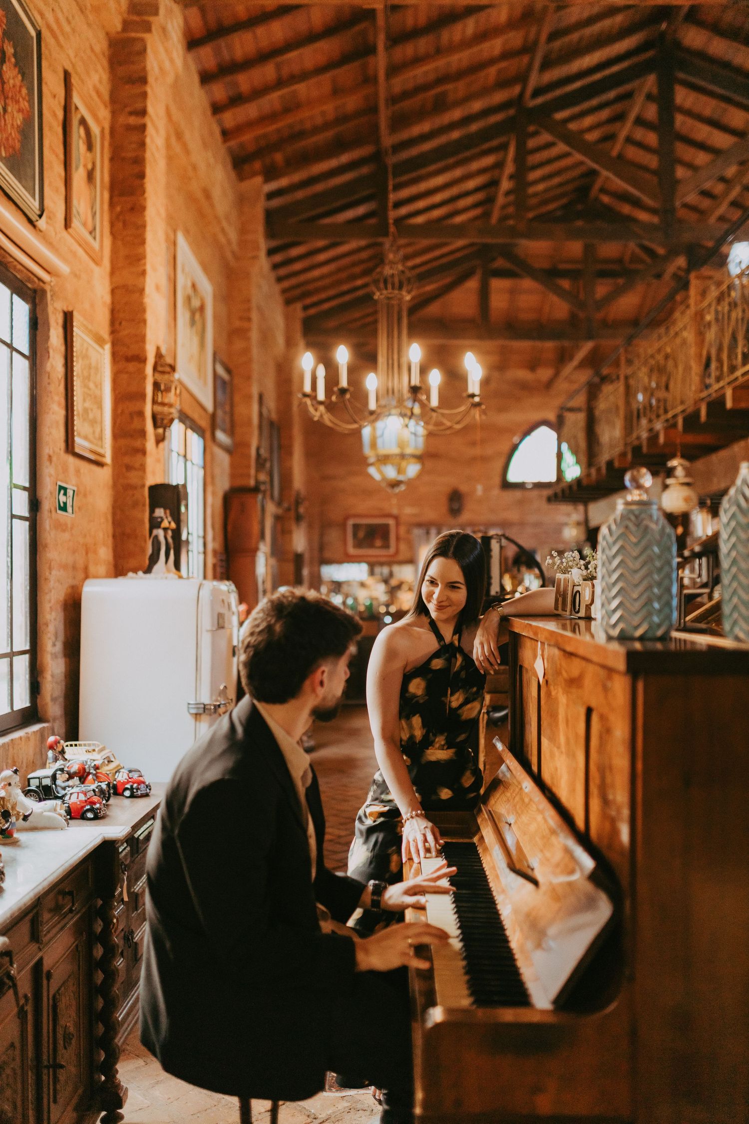 A man plays piano for a smiling woman in a rustic brick interior with chandelier lighting.