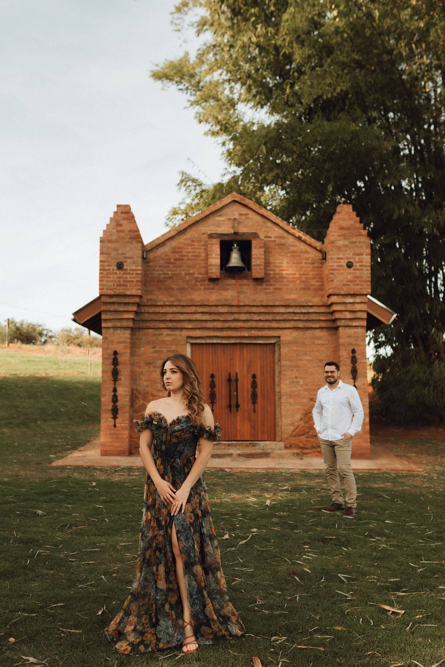 Couple poses before a rustic brick chapel surrounded by lush greenery, woman in floral gown, man in white shirt.