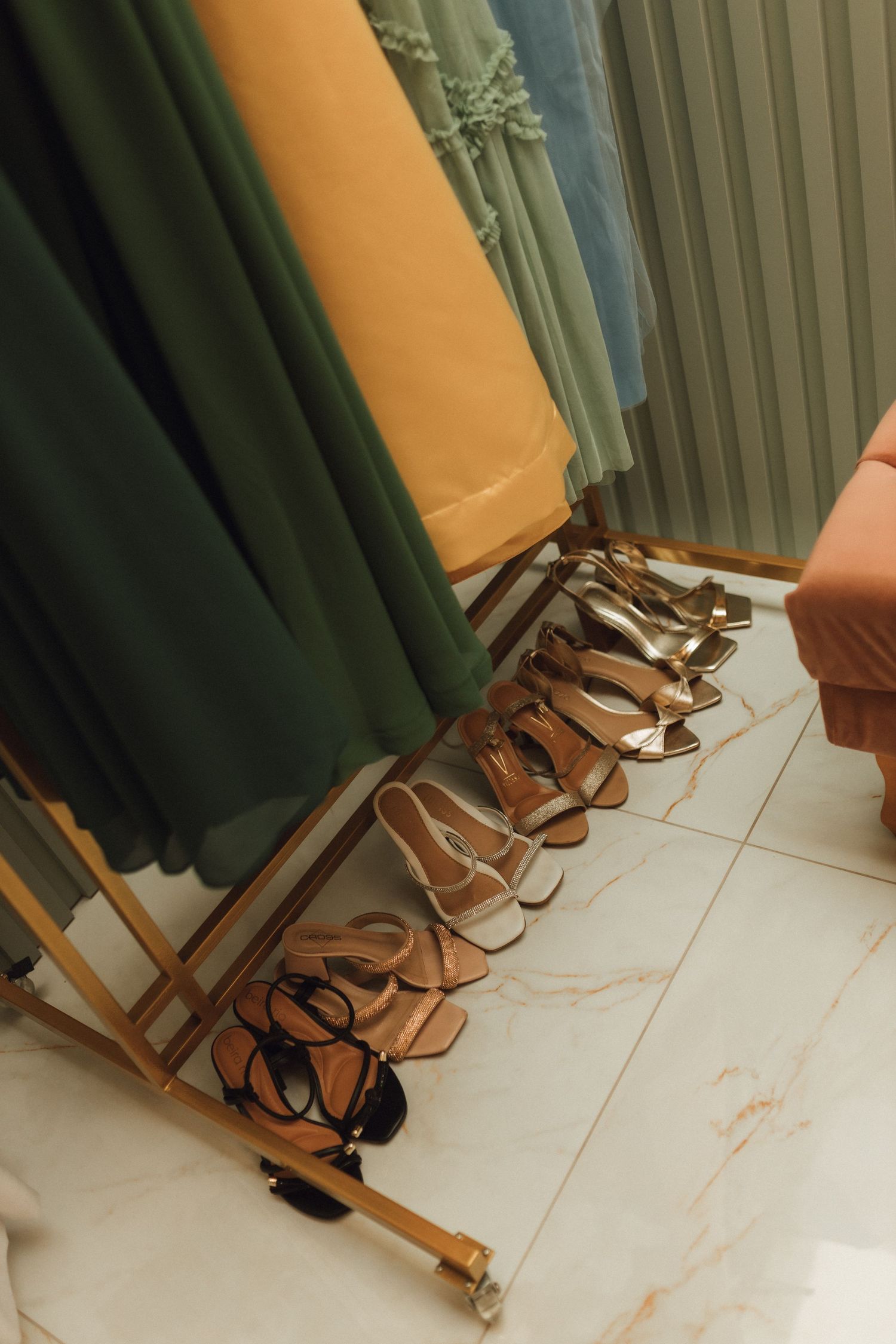 Gold heeled sandals lined up beneath colorful bridesmaid dresses hanging on a clothing rack indoors.