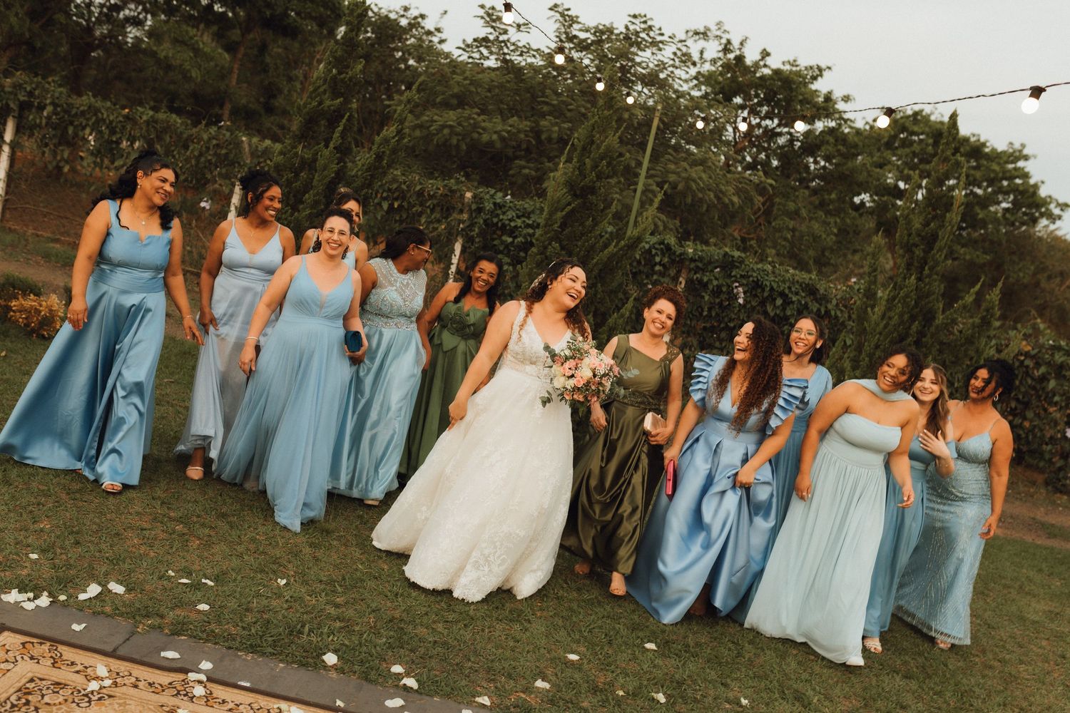 Bride in white gown poses with bridesmaids in blue dresses on green lawn at outdoor wedding celebration.