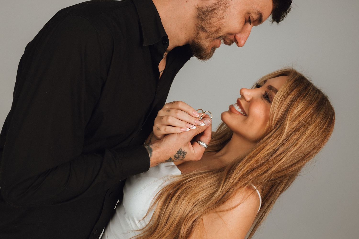 A smiling couple holds hands and gazes at each other, the man in black shirt and woman in white dress, against a gray background.