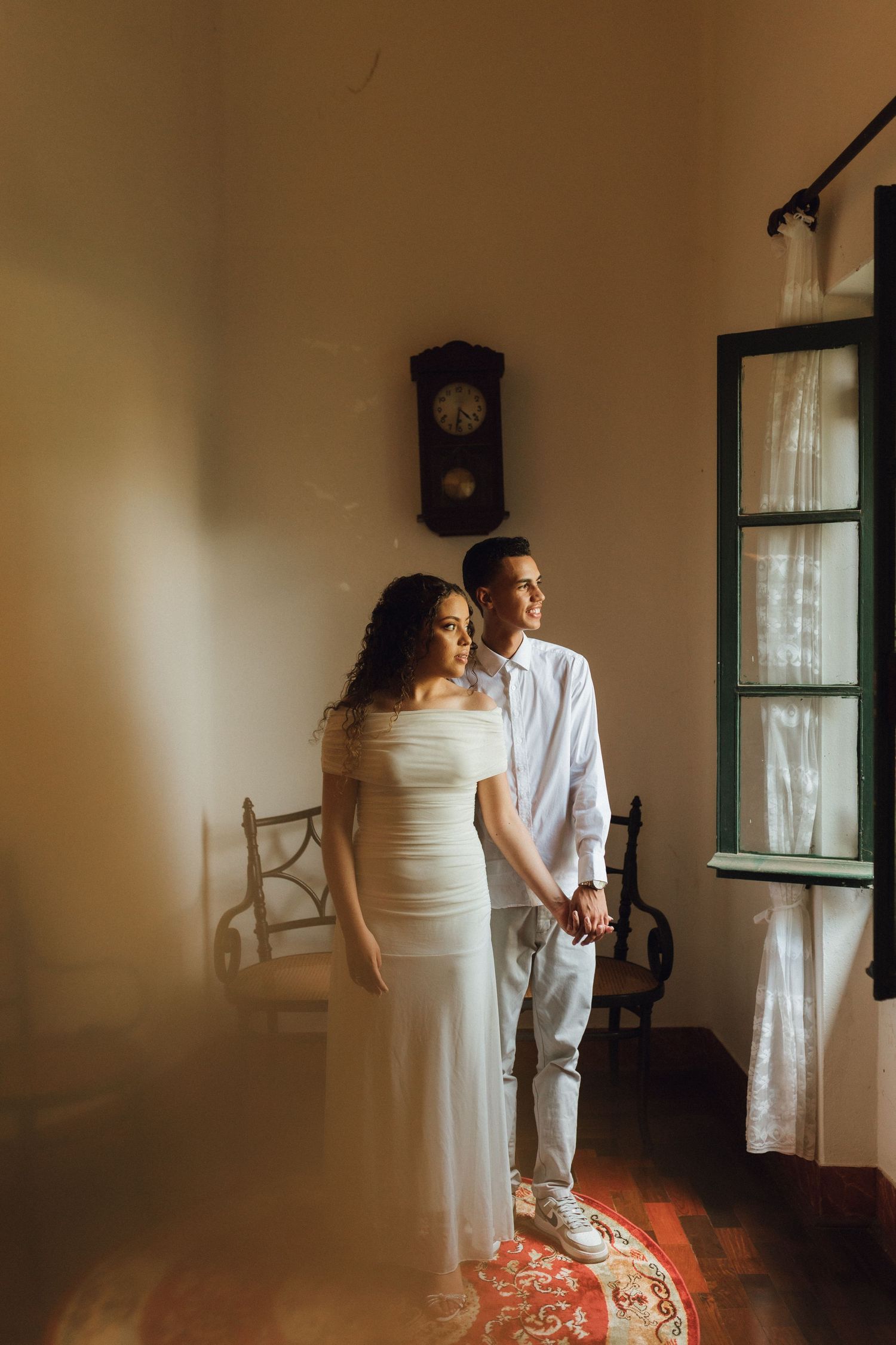 Couple in white outfits holding hands by a sunlit window with vintage clock and antique chairs in elegant room.