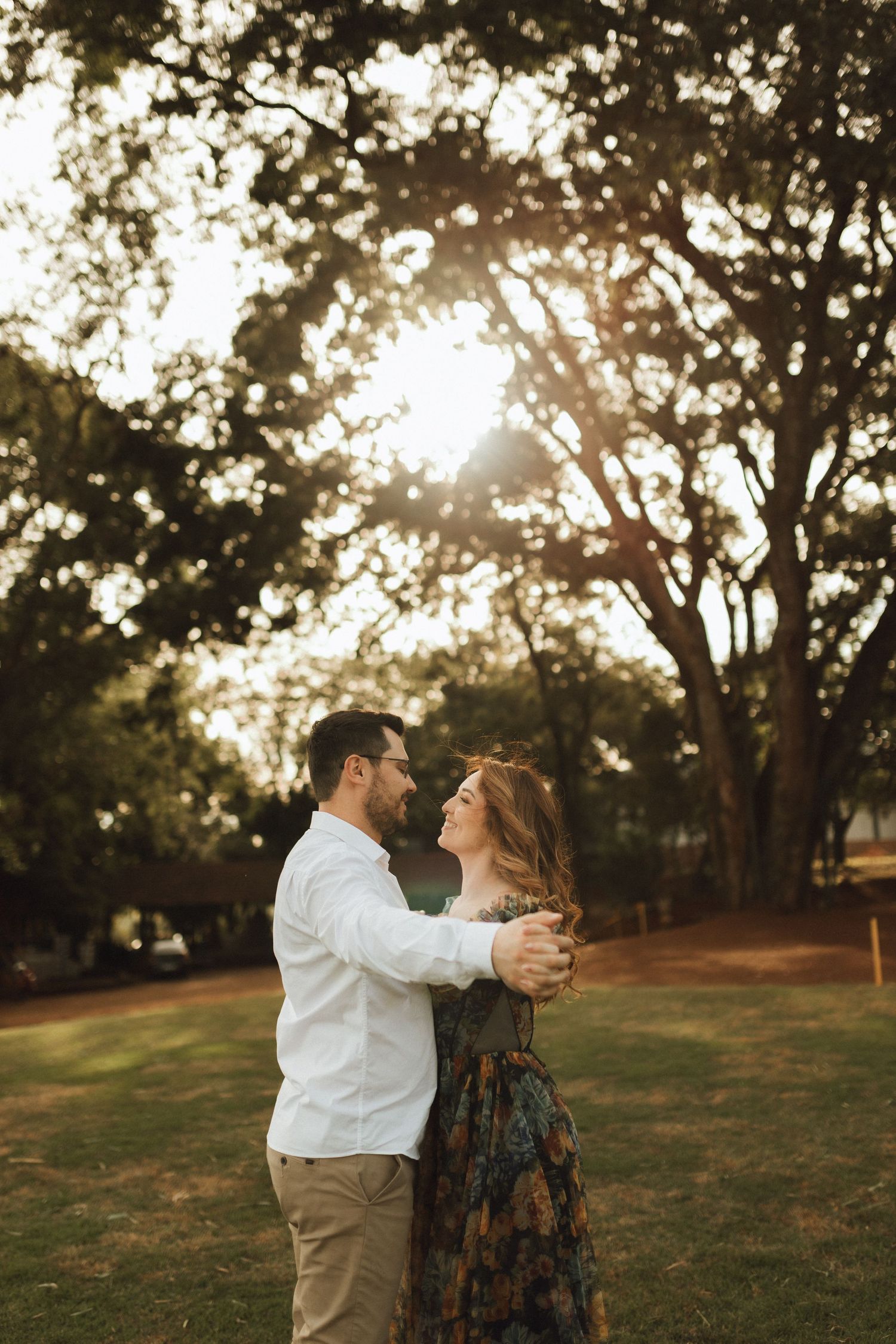 Couple embracing at golden hour in a lush green park, sun rays filtering through tall trees behind them.