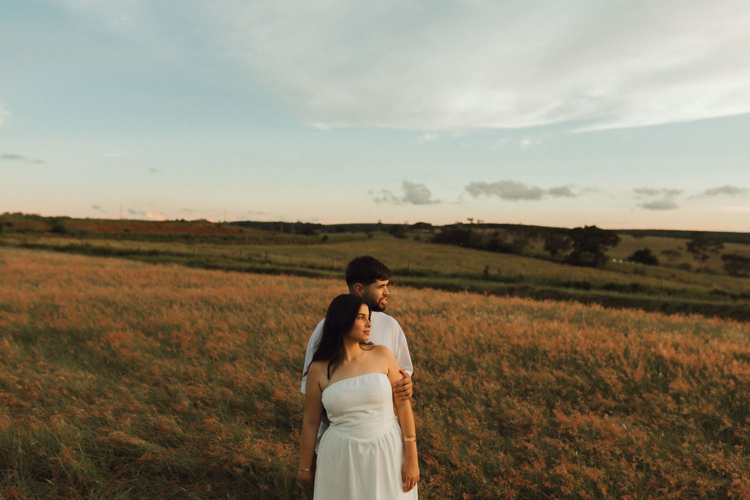 Couple standing together in an open golden field at sunset, gazing into the distance.