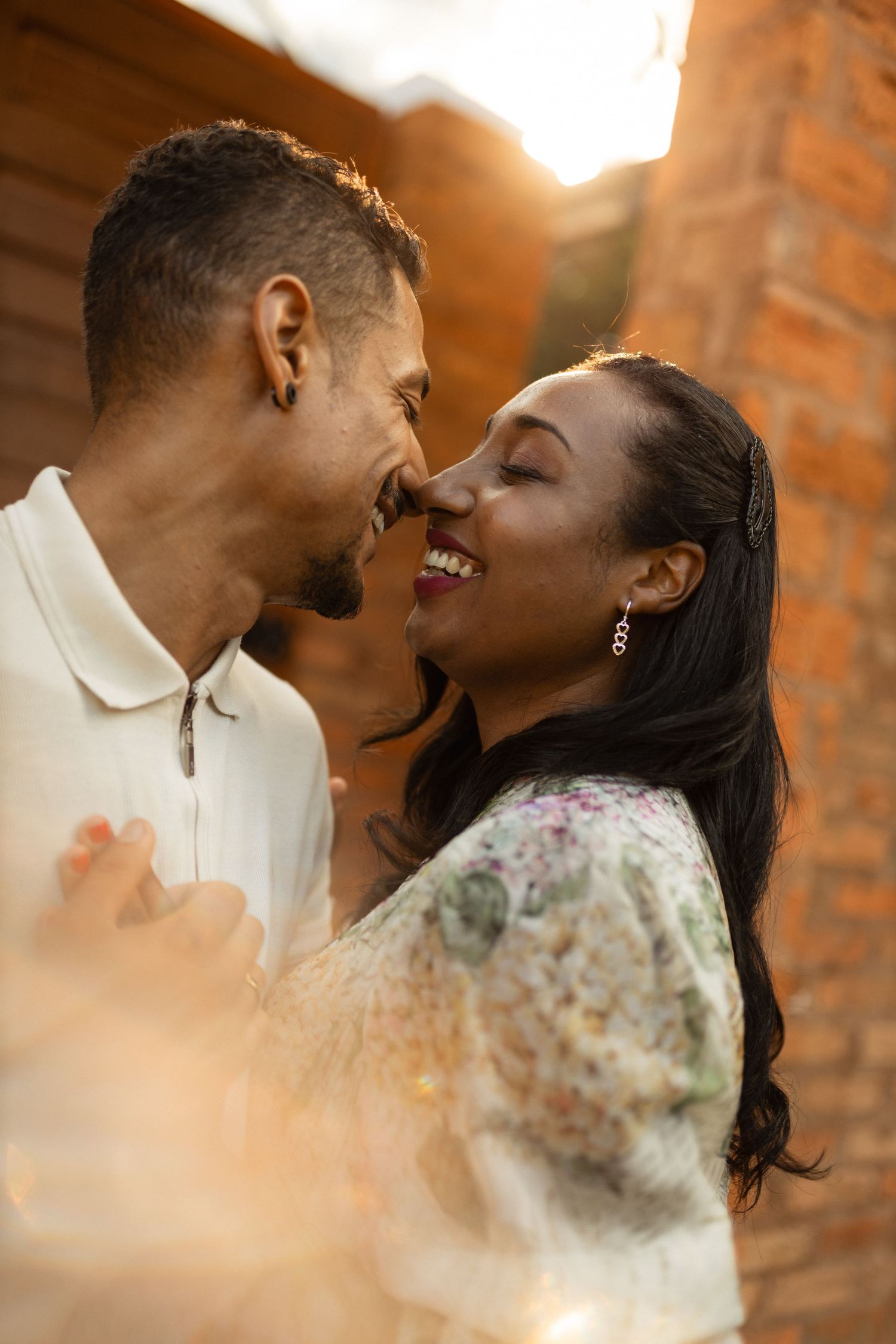 Romantic couple nearly kissing in warm golden sunset light by brick wall.