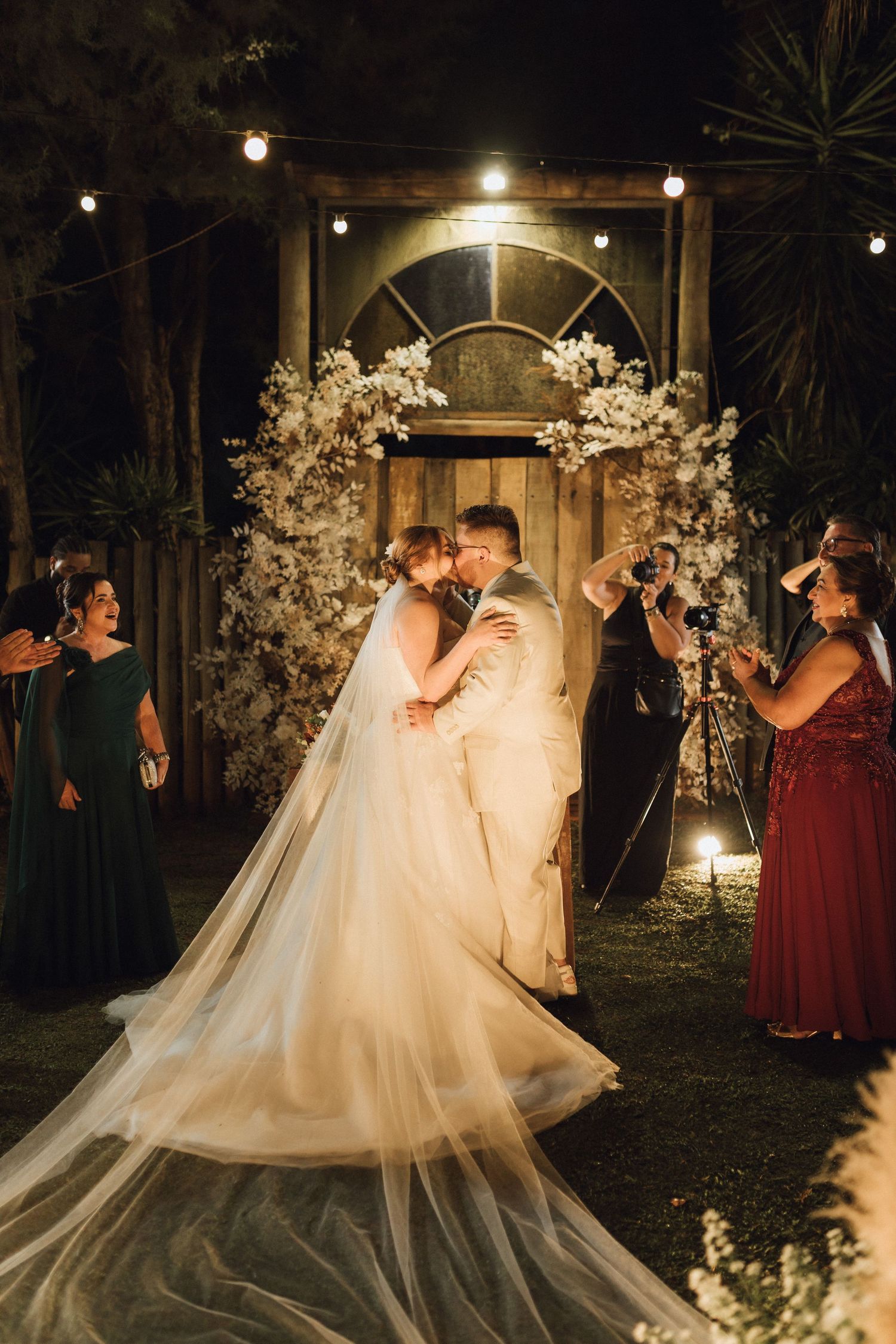 Newlyweds share first kiss under floral arch with string lights at outdoor night wedding.