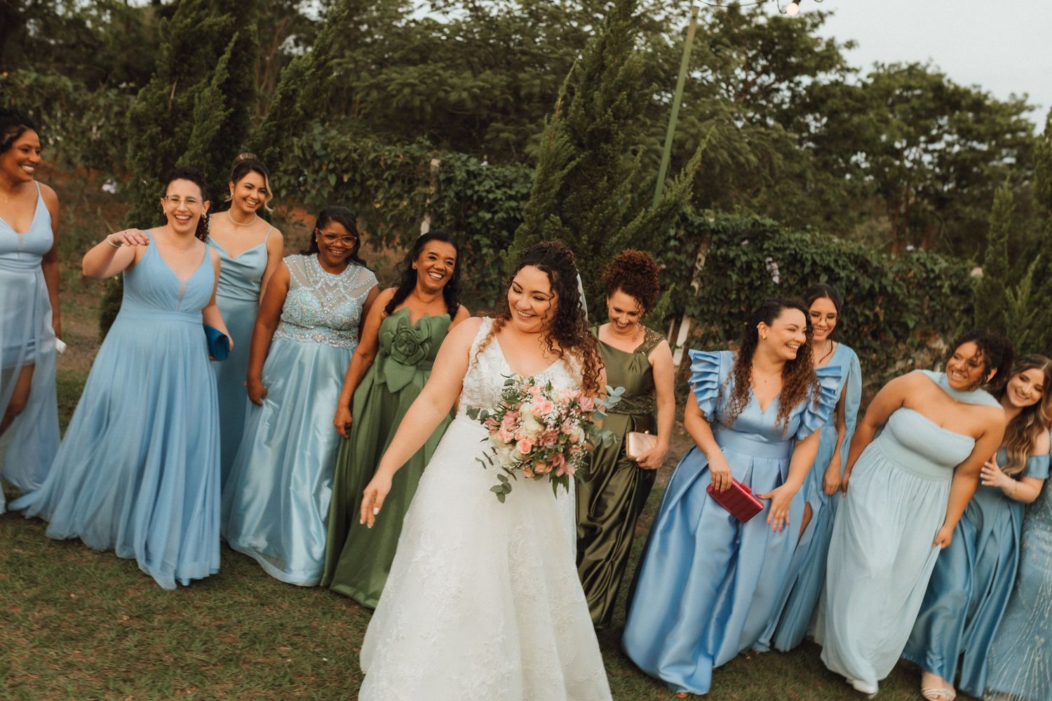 Bride walks joyfully with bridesmaids in blue and green dresses at an outdoor wedding celebration.