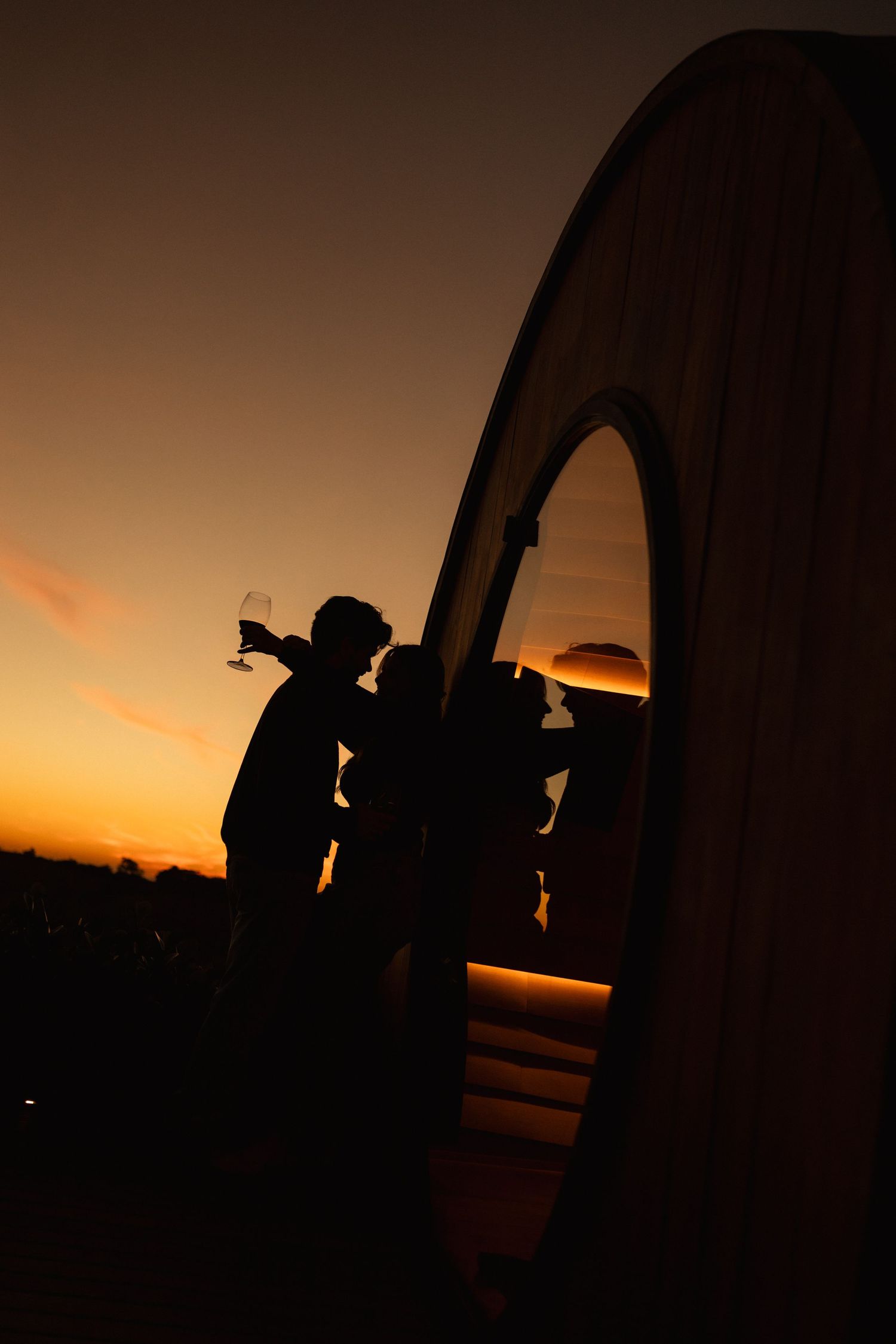 Silhouette of two people holding wine glasses at sunset beside a large circular architectural structure.