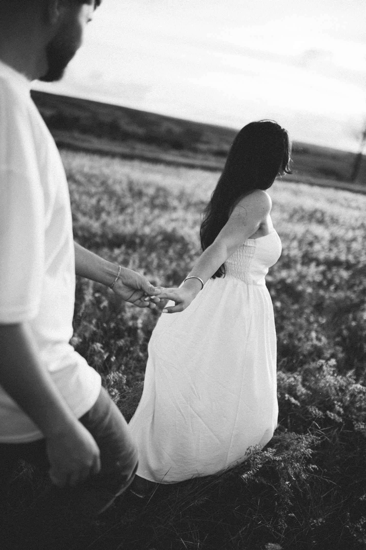 Black and white photo of a couple holding hands in a field, woman in white dress with long dark hair.