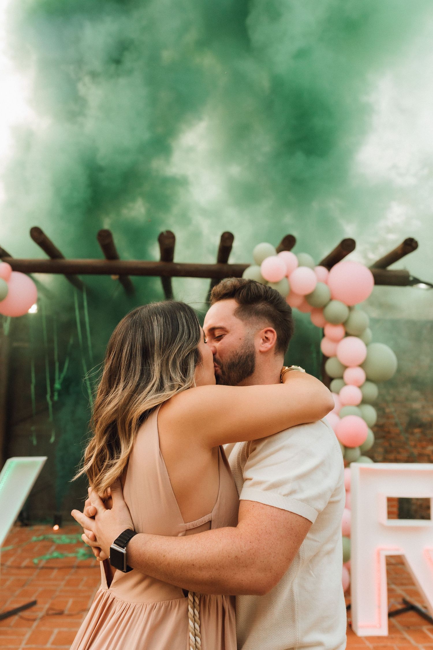 Couple kissing and embracing at a gender reveal party with green smoke, pink and sage balloon arch backdrop.