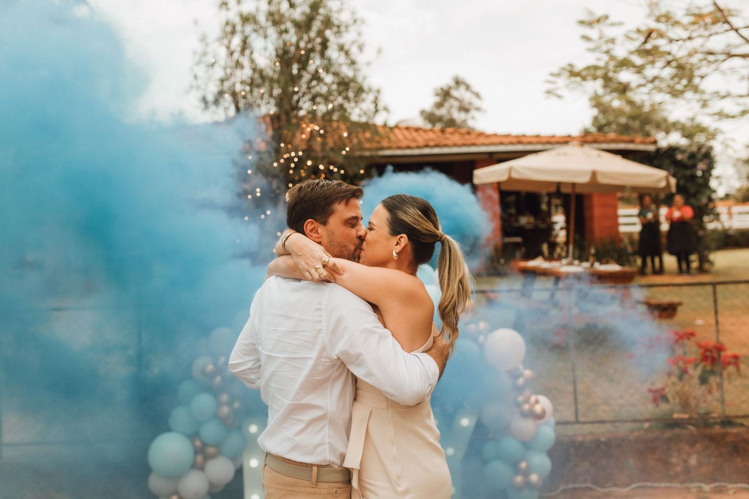 Couple kissing surrounded by blue smoke bombs at outdoor gender reveal party with balloon decorations.