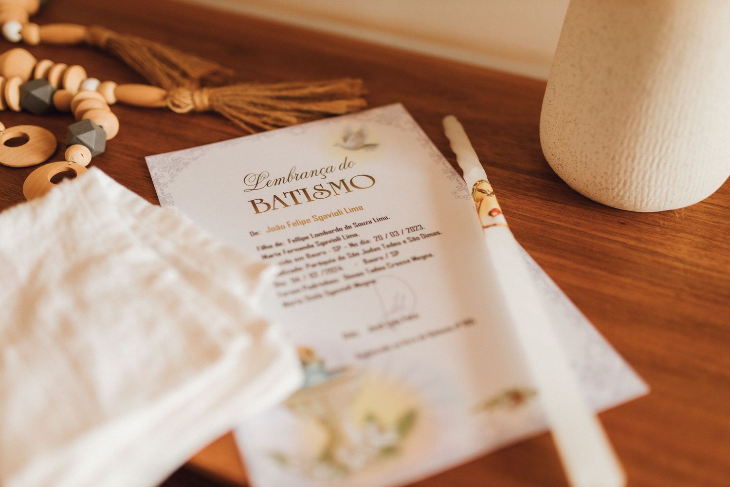 Baptism invitation card with candle, rosary beads, and white fabric on wooden table for christening ceremony.