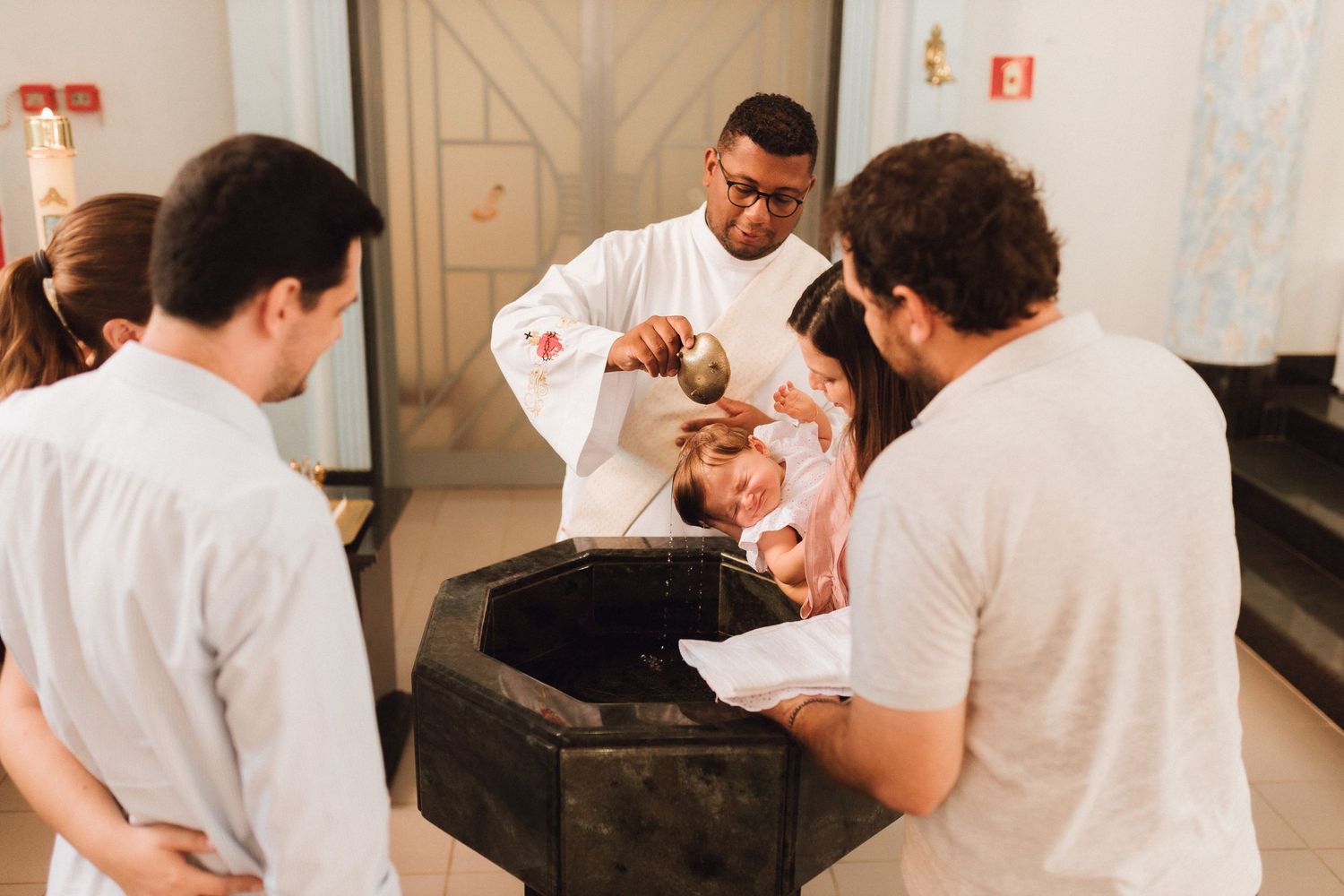 Priest performing a baptism ceremony, pouring holy water over an infant at a baptismal font.