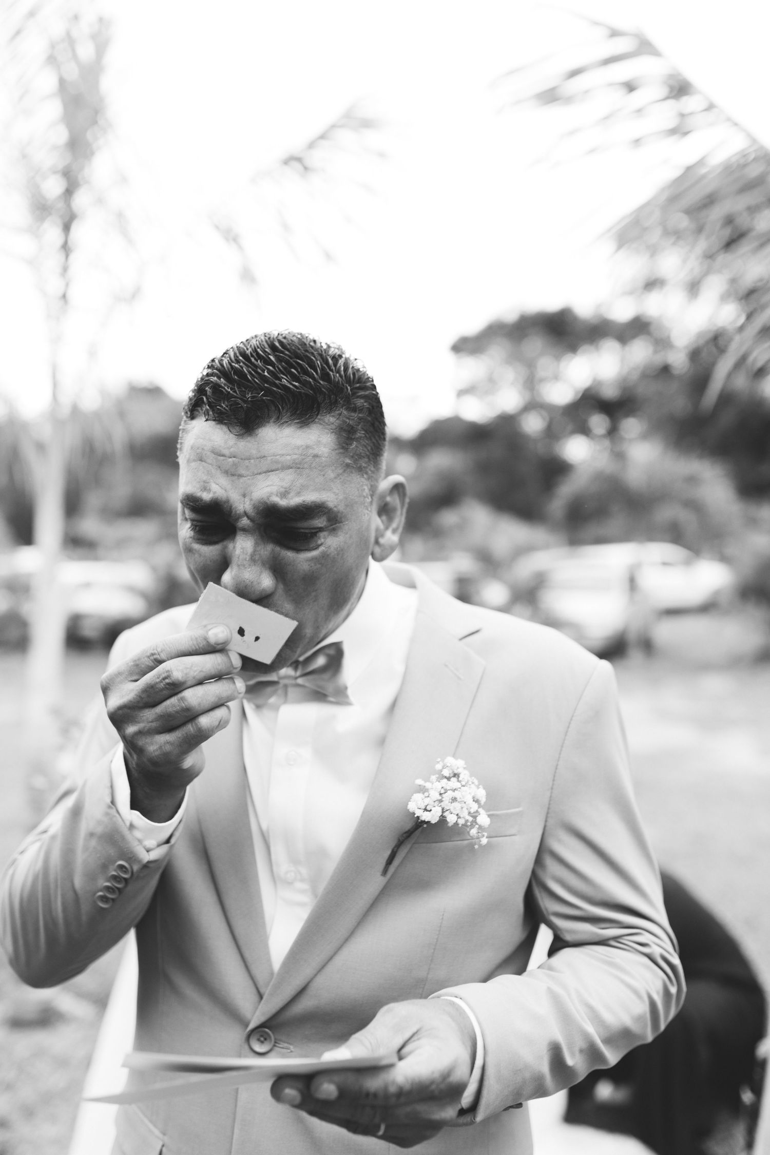 Emotional groom in gray suit wipes tears while reading vows card at outdoor wedding ceremony.