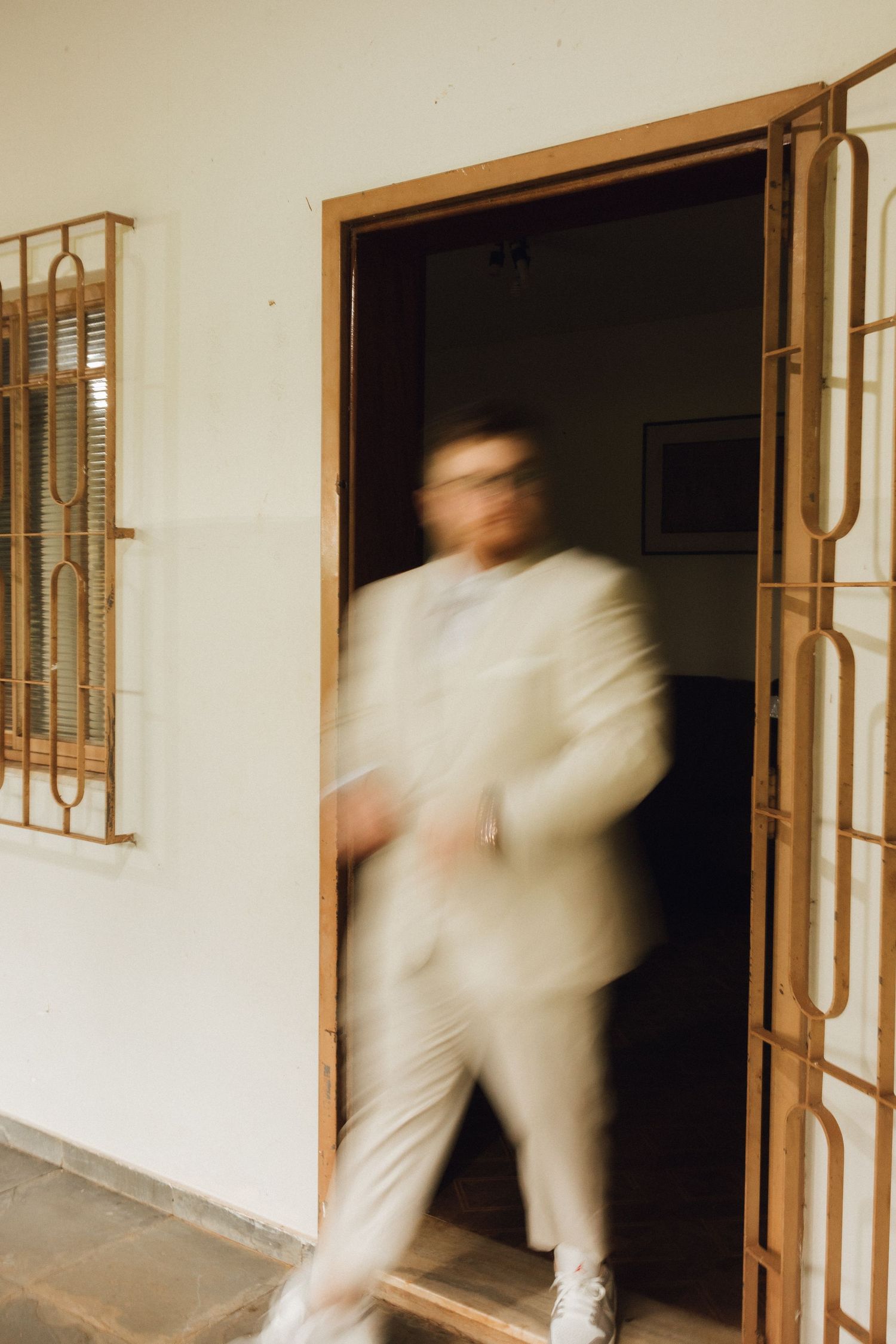 Blurred motion shot of groom in cream suit walking through wooden doorway at wedding venue.