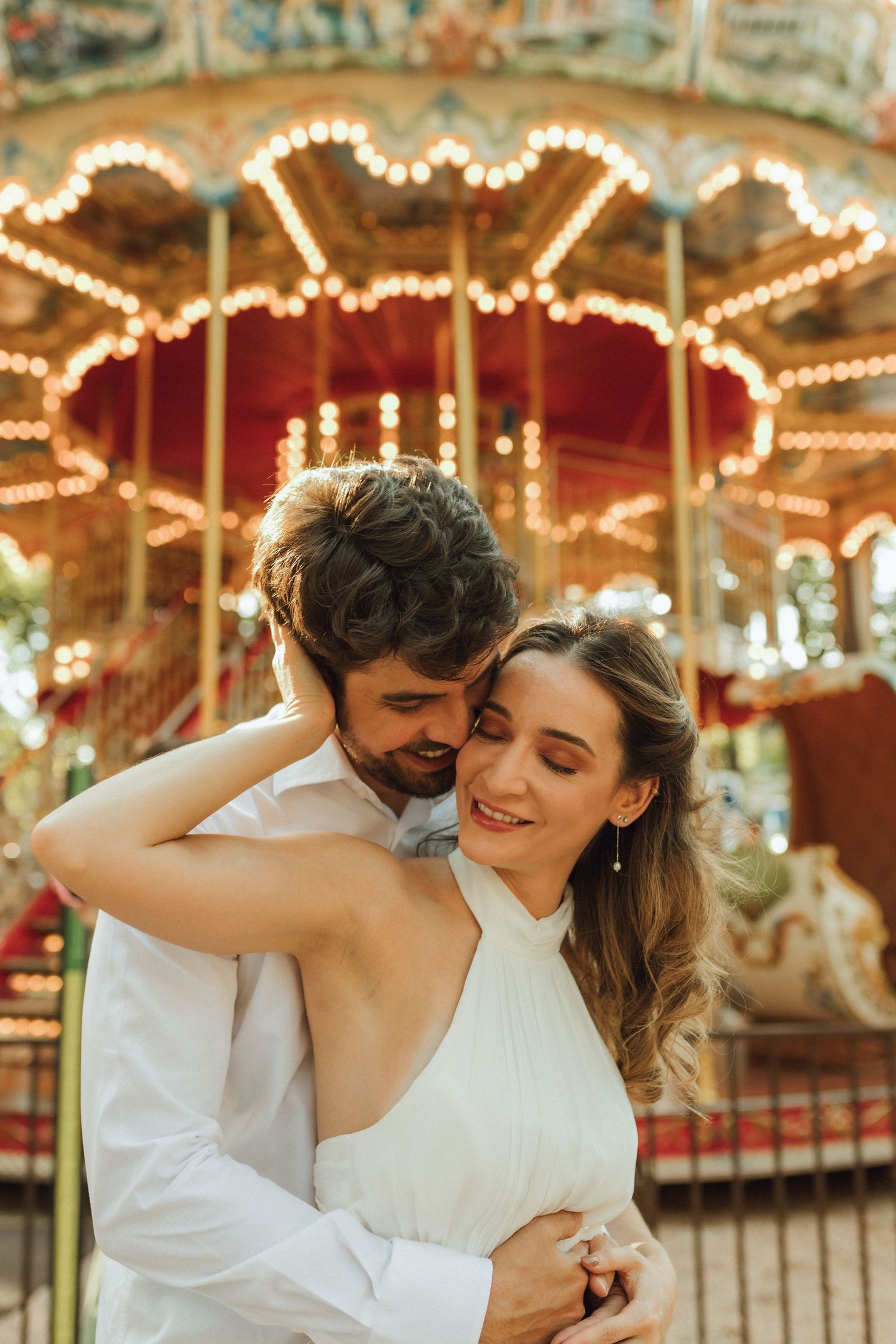 Romantic couple embracing and smiling in front of illuminated carousel at amusement park.
