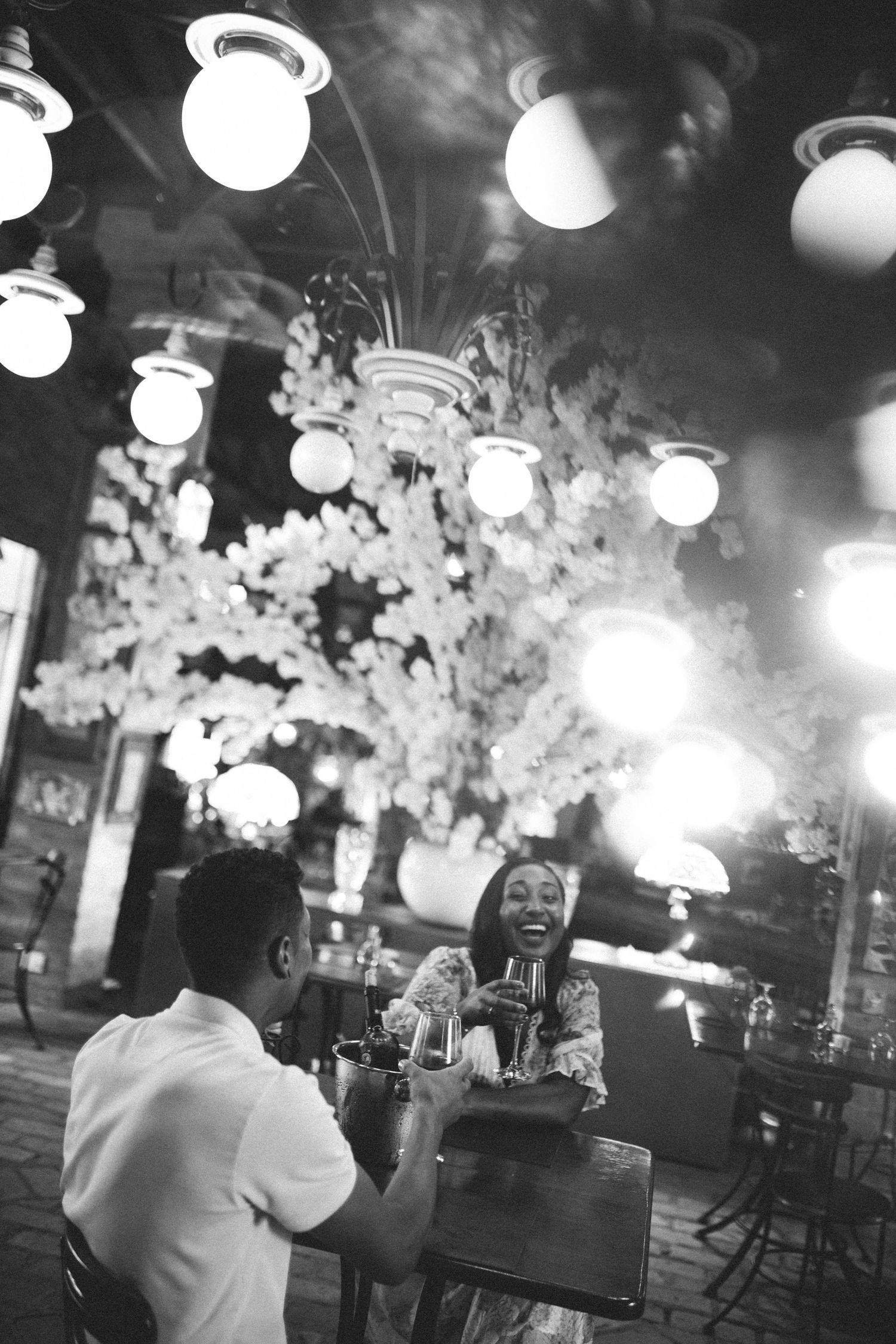 Couple laughs and clinks glasses at candlelit table beneath glowing lights at elegant night reception.