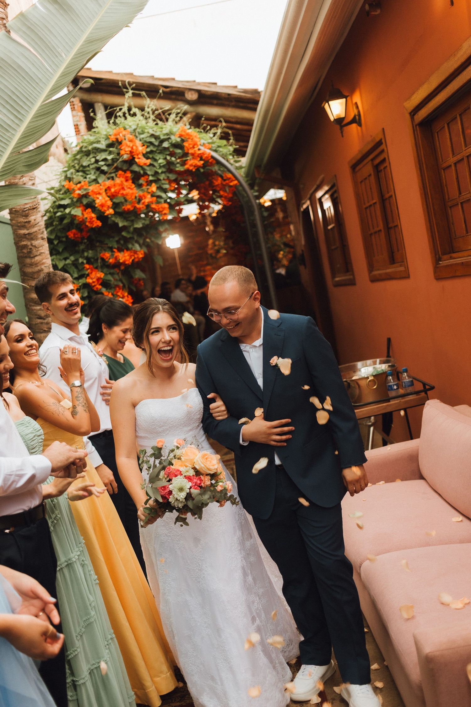 Joyful newlyweds exit ceremony as guests shower them with flower petals at colorful outdoor venue.