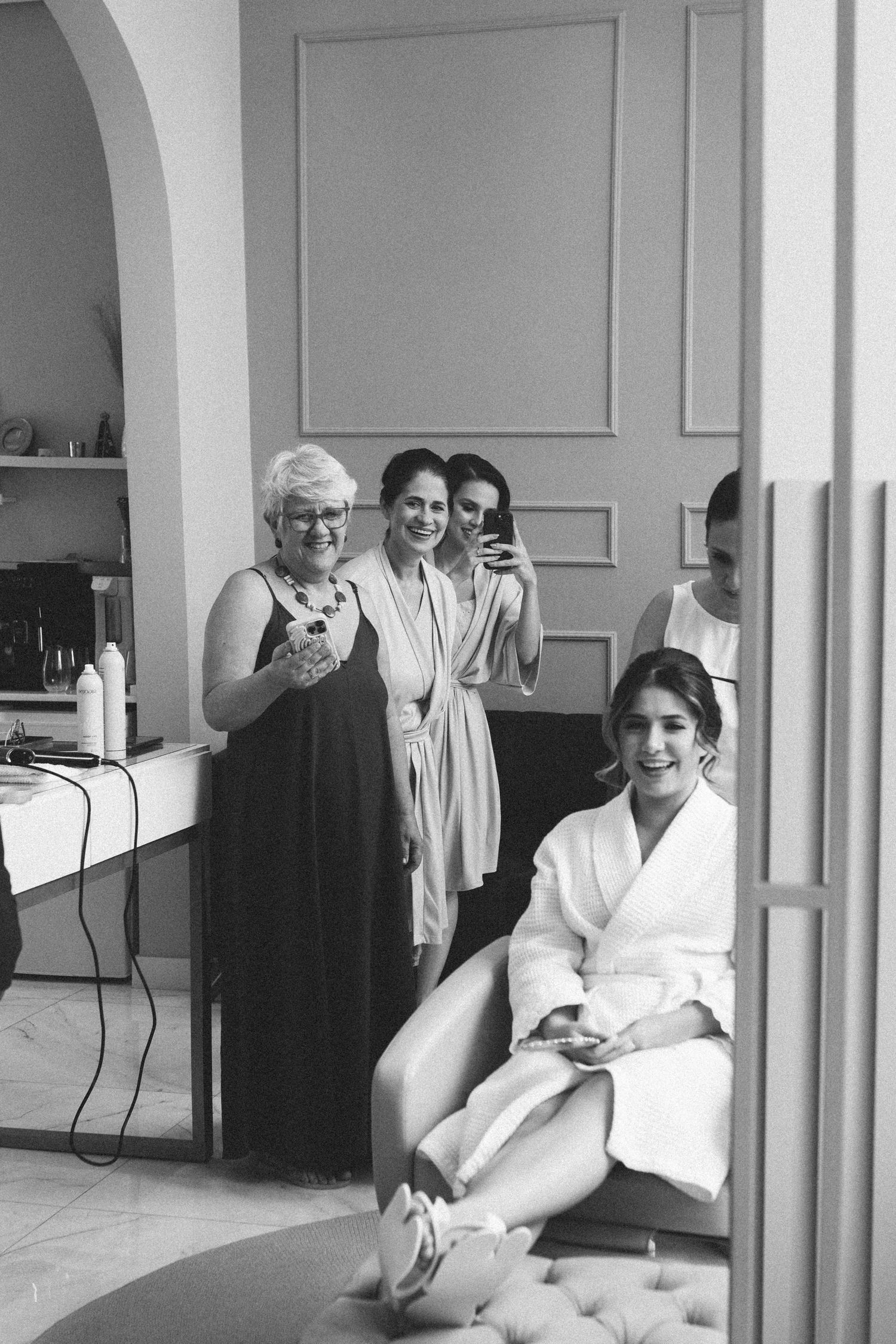 Black and white mirror selfie of bride smiling in robe with bridesmaids and mother during wedding preparations.
