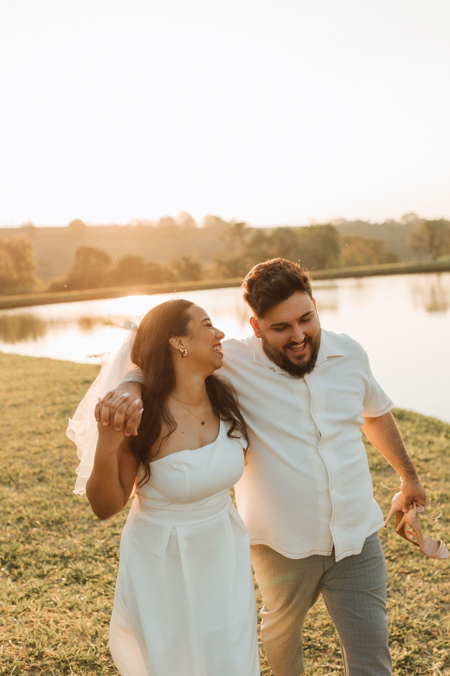 Happy newlywed couple walking outdoors by a lake at golden hour, bride in white dress and veil, groom in white shirt.